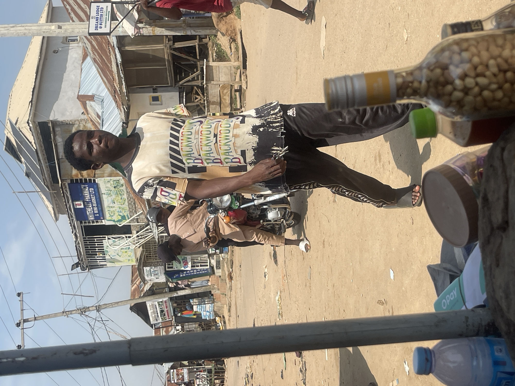 A full-body shot captures a young Black man standing outdoors on a dusty, sun-drenched street in Abuja, Nigeria. He wears a cream-colored t-shirt with a repeating "BACKWOODS" graphic print and black track pants with white stripes along the sides. The man holds a set of keys in his left hand and glances towards the camera with a neutral expression. To his right, a clear glass bottle filled with small, light-colored seeds sits prominently, and in the background, a motorbike is visible.
The scene appears to be a busy commercial area, with various shops and stalls lining the street. A sign for "WHITEHEART PHARMACY & MINI-MARKET LTD" is visible on the left, and another sign reads "DREAMS LODGE & SUITES LTD". Overhead, power lines crisscross the sky, suggesting an urban environment. The lighting and shadows indicate it is daytime, likely mid-morning or afternoon, with bright sunlight. The ground is covered in dirt and scattered debris, typical of a street in this region. The overall impression is one of everyday life in a Nigerian city, with people engaged in various activities and commerce.