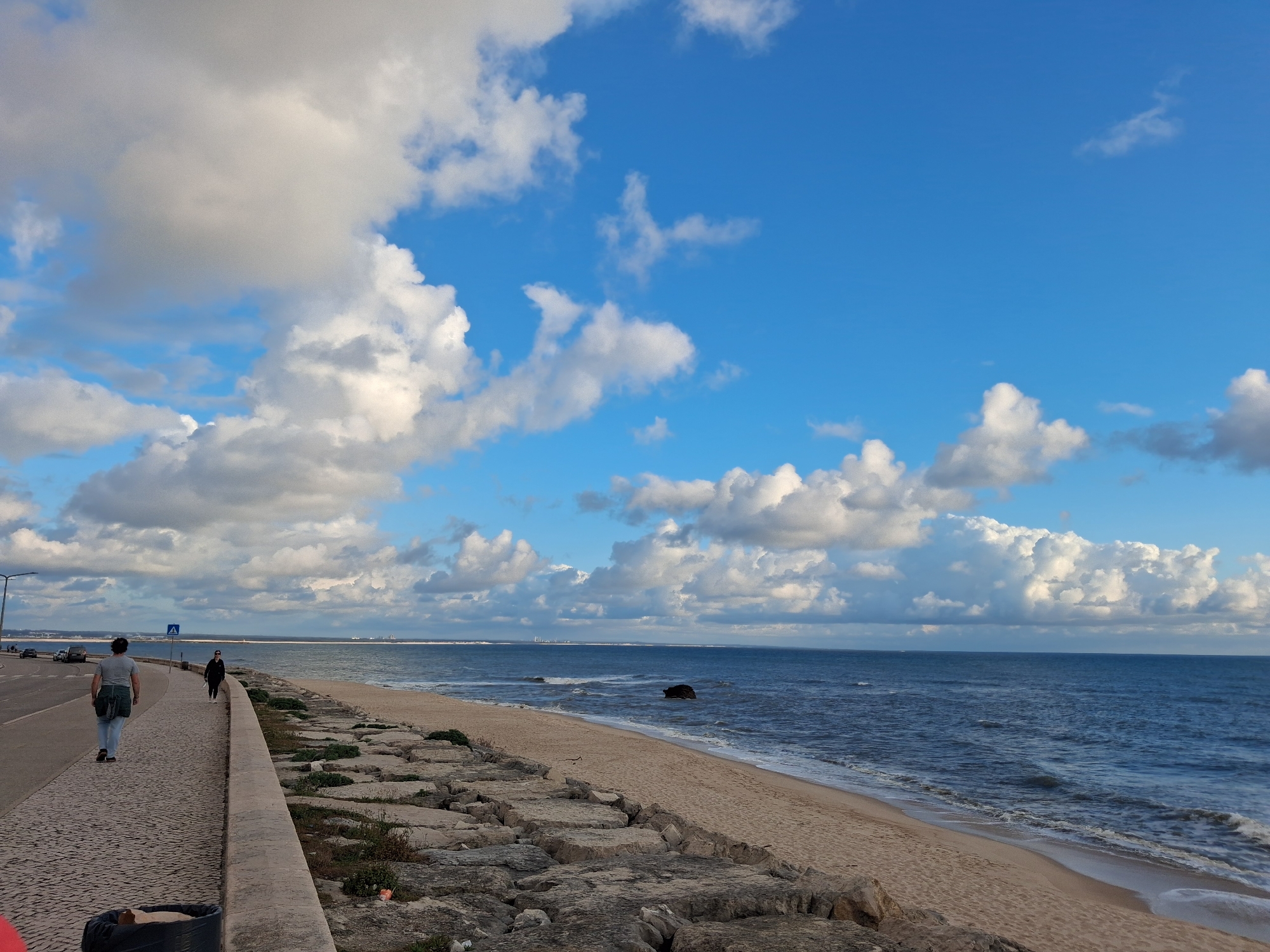 The image shows a coastal scene in Figueira da Foz, Portugal. A paved walkway runs parallel to a sandy beach, separated by a low concrete wall reinforced with large rocks. Two people are walking along the walkway, one further away and another closer to the camera. Cars are parked on the roadside along the walkway. The ocean stretches to the horizon, with a few visible waves and a large rock formation near the shore. The sky is predominantly blue, with numerous white and gray cumulus clouds scattered throughout. The overall lighting suggests a daytime scene with diffused sunlight.