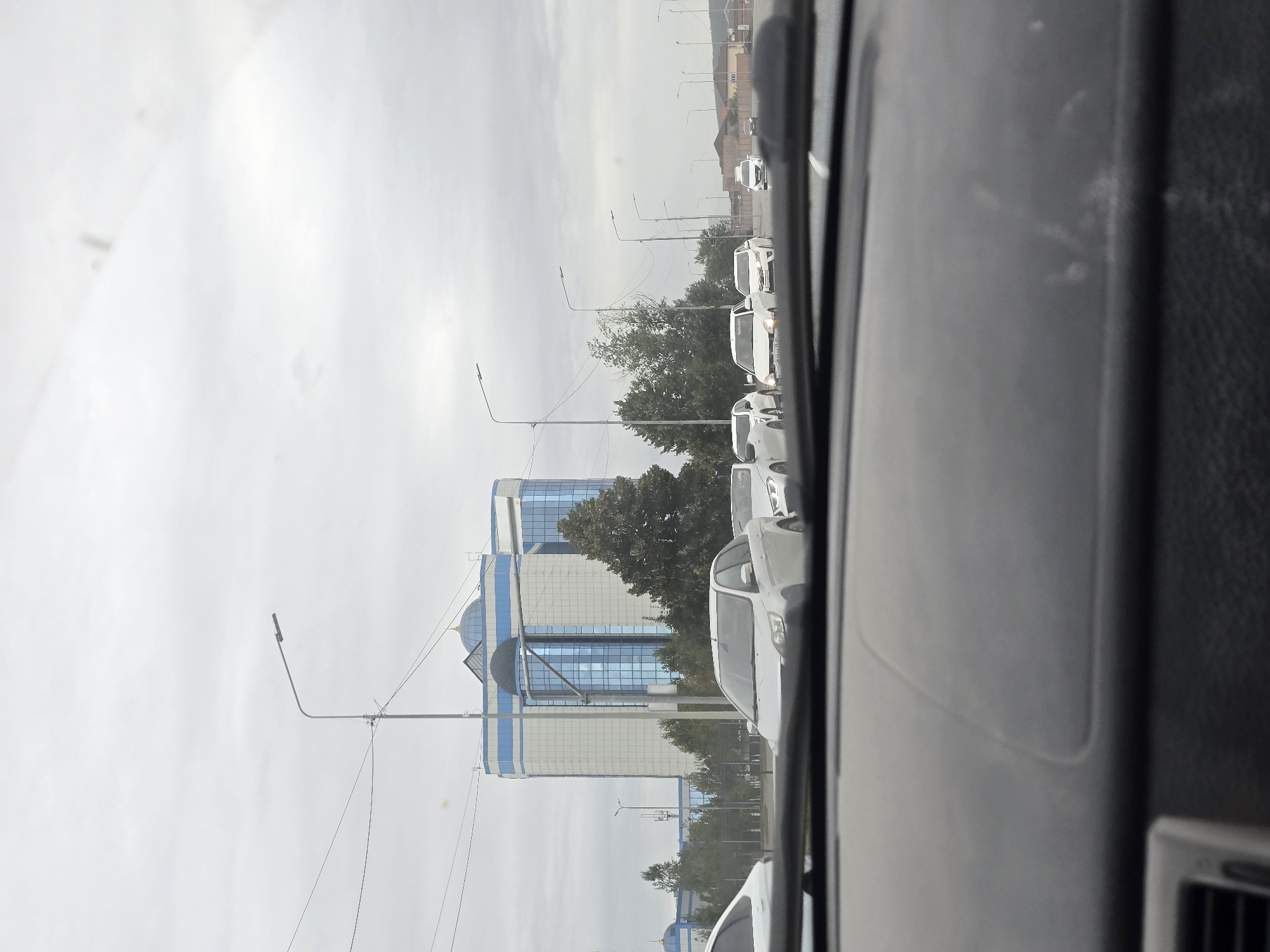 This is a street-level, medium shot, eye-level, horizontal, full-color photograph taken from inside a vehicle. The perspective is through the windshield, which shows the dashboard in the foreground. The scene depicts a cloudy day with a wide road with multiple lanes of traffic. Several white cars are visible, some moving and some stationary. In the background, there is a large, modern, multi-story building with a blue and white facade and a prominent blue dome on top. Trees line the street, and streetlights are visible. The image appears to be a candid shot of traffic in an urban environment, likely in Grozny, Russia, given the architectural style of the building and the general urban setting.