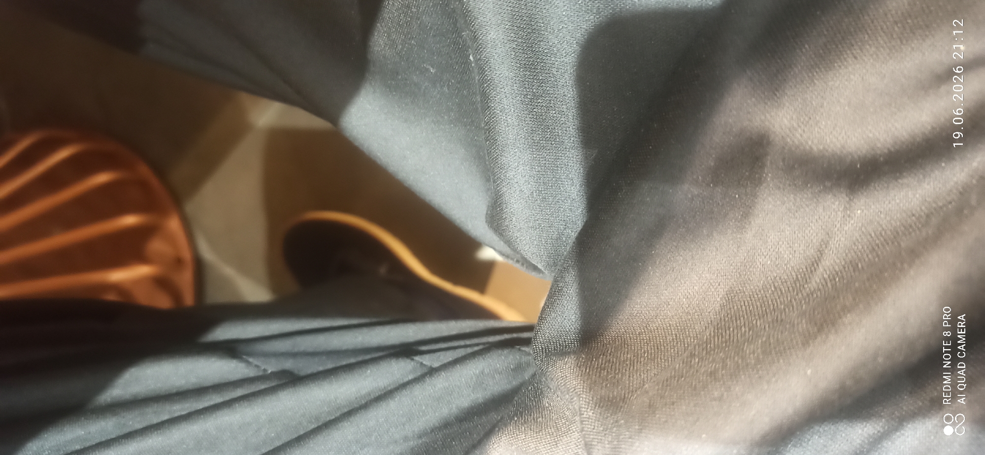 The image displays a close-up view of dark fabric, likely clothing, with a textured weave visible. The fabric is creased and folded, suggesting it is being worn or is draped. In the background, out of focus, is an orange object with a ribbed surface, possibly a piece of furniture or a decorative item. There is also a glimpse of a brown object, possibly a shoe or a sandal, with a light-colored sole. The lighting creates shadows and highlights on the fabric, indicating an indoor setting.
The text overlay in the bottom right corner provides a timestamp: "19.06.2026 21:12". Below this, it reads "REDMI NOTE 8 PRO AI QUAD CAMERA", indicating the device used to capture the image and that it was taken at night. The overall impression is a casual, candid shot, possibly taken by someone sitting or lying down, with the focus on the texture and folds of the fabric. There are no people or clear signs of a specific activity or event. The image is taken from a low-angle perspective, looking upwards at the fabric.
