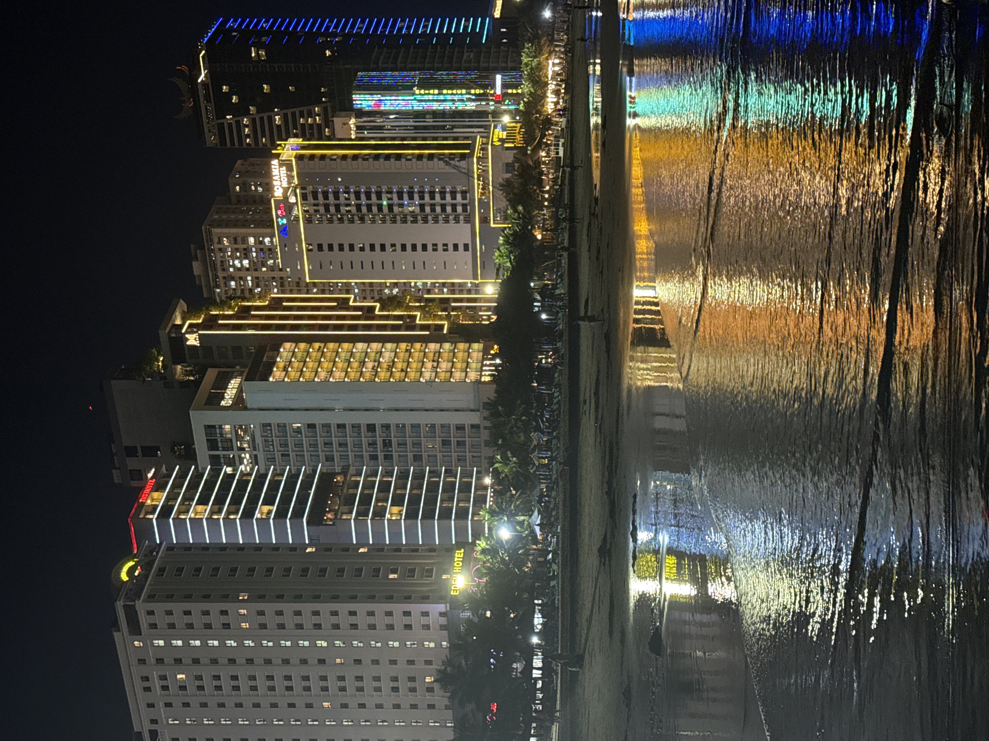 This media file depicts a nighttime scene of a beachside city in Đà Nẵng, Vietnam. The image is vertically oriented, showing a row of illuminated hotels lining a beach. The hotels are modern structures with numerous windows and are adorned with bright, colorful lights, primarily white and yellow, outlining their architectural features. Some buildings display neon signs, including one that reads "EDEN HOTEL" in red lettering.
The beach itself is visible in the foreground, with gentle waves reflecting the vibrant lights of the hotels. The water's surface is textured, creating a mesmerizing play of light and color. Scattered on the beach are a few palm trees, silhouetted against the bright hotel facades, and a few small figures of people can be discerned walking along the shore.
The time of day is clearly night, with the sky being a deep, dark black, emphasizing the artificial lights of the city. The weather appears calm, as indicated by the smooth reflections on the water. The overall atmosphere is lively and vibrant, characteristic of a popular tourist destination at night. The combination of modern architecture, beachside setting, and colorful illumination suggests a festive and engaging environment.