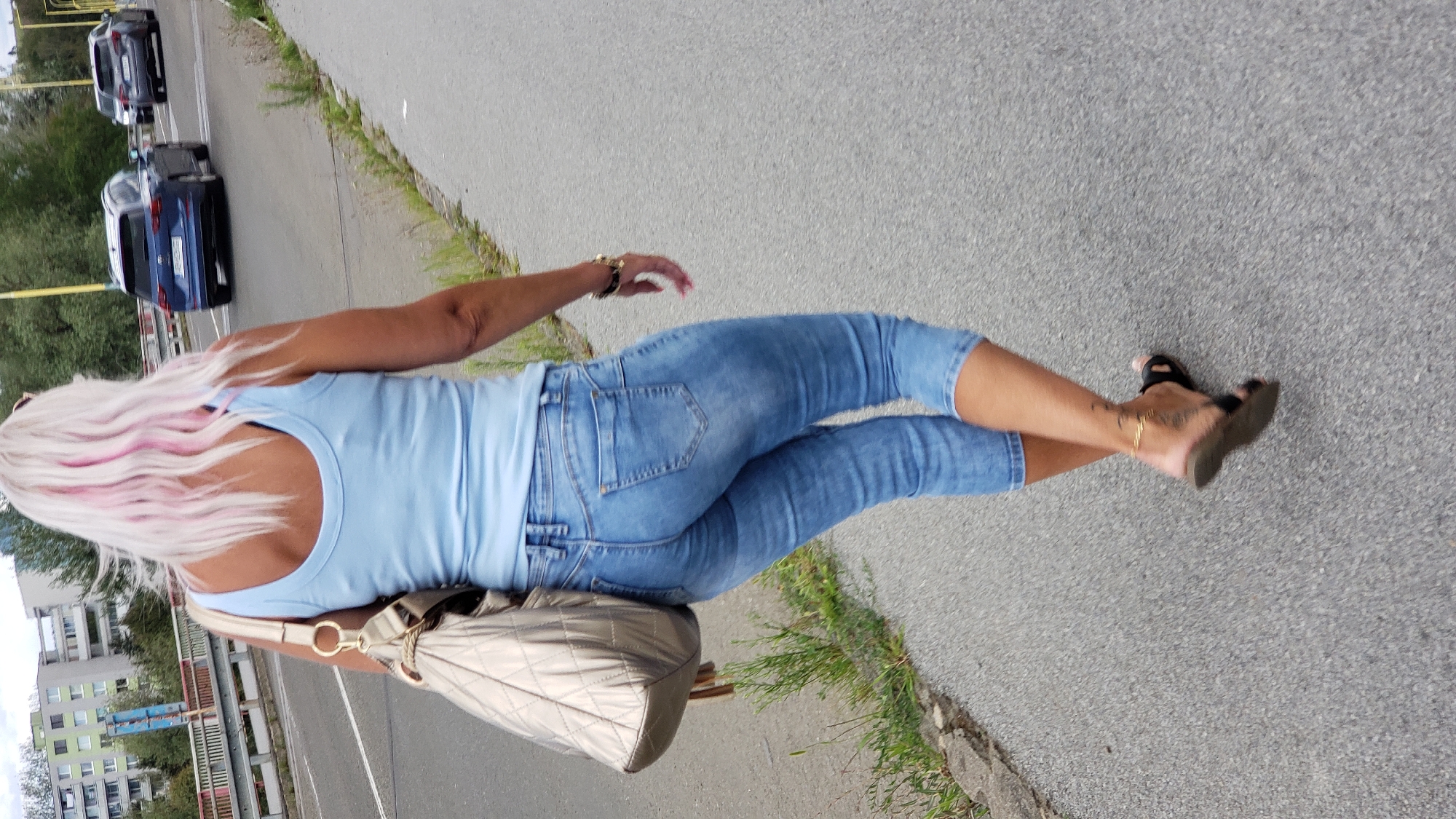 A woman with blonde hair with pink highlights, wearing a light blue tank top and blue capri jeans, walks on a paved sidewalk next to a road in Prešov, Slovakia. She carries a gold quilted shoulder bag and wears black sandals. A tattoo is visible on her left ankle. In the background, cars are driving on the road, and buildings and trees are visible.