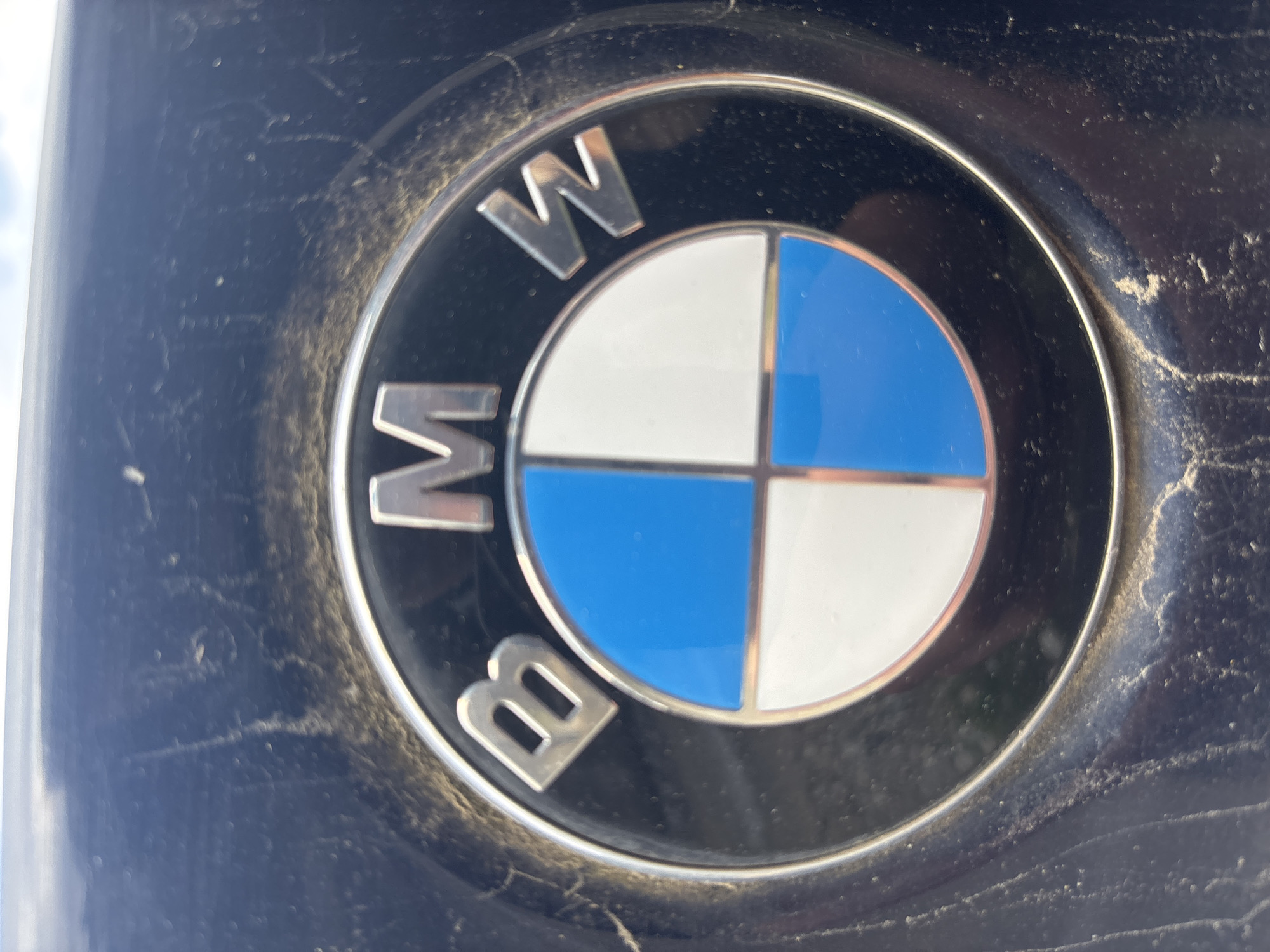 A close-up, slightly dusty, BMW logo is depicted. The logo features the letters "BMW" around the perimeter of a circle divided into four quadrants of alternating blue and white. The background surface appears to be dark and slightly worn. The location of the logo in Zlatograd, Bulgaria is not evident from the image.