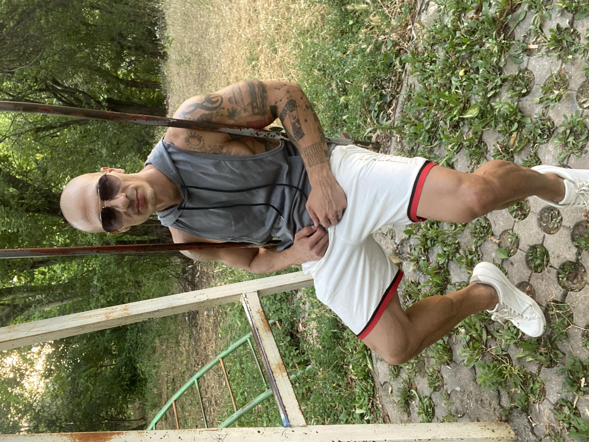 A bald, fair-skinned man with sunglasses is sitting on a swing set. He is wearing a grey sleeveless hooded shirt and white shorts with red trim. The man has tattoos on both arms. He is positioned in the center of the frame, with the swing set structure on either side. The background is a natural setting with trees and overgrown vegetation. The ground is paved with interlocking concrete blocks interspersed with grass. A green metal ladder is visible on the left side of the frame, attached to the swing set structure. The scene appears to be in an outdoor recreational area. The image was taken in Pleven, Bulgaria.
