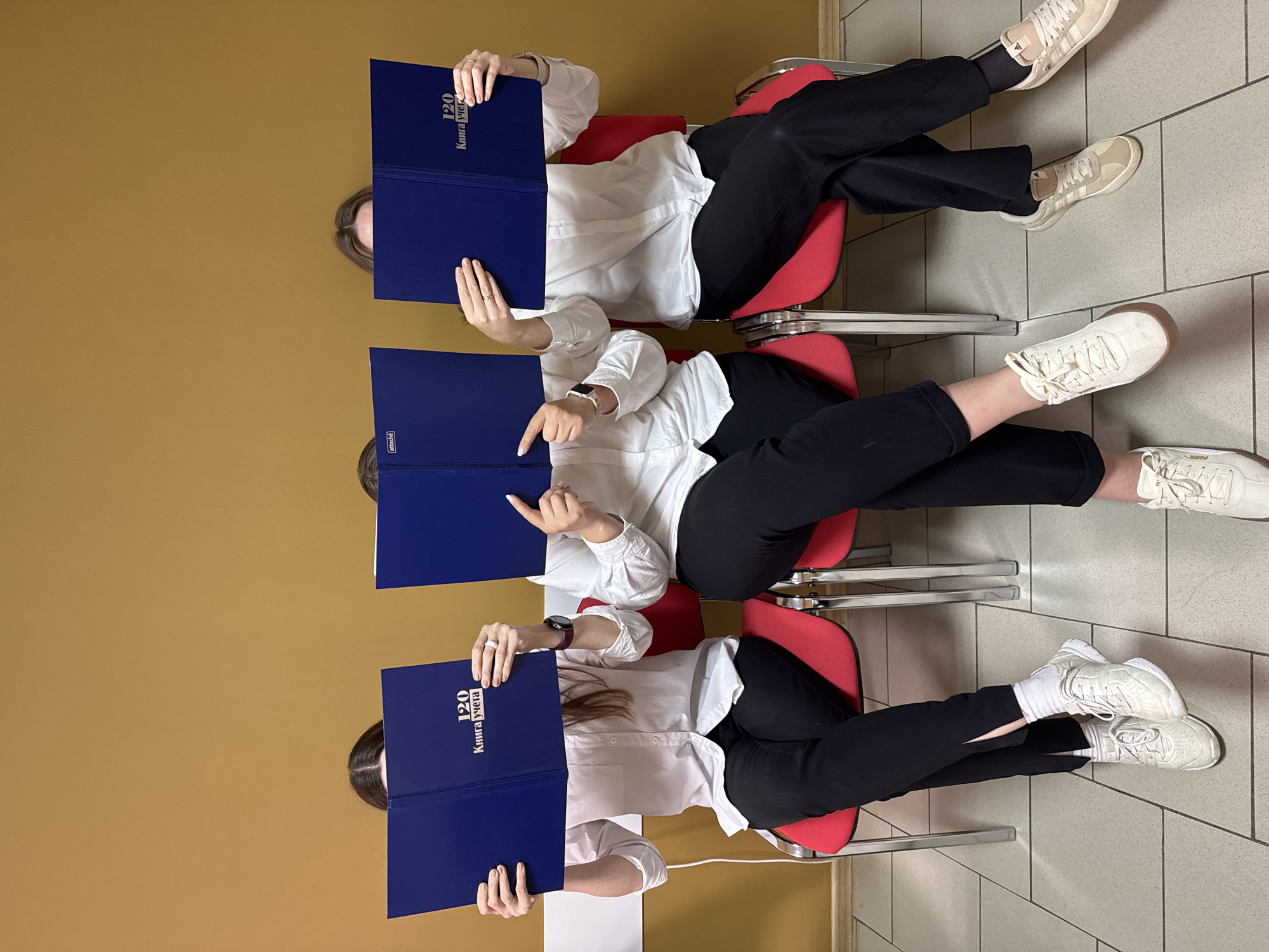 Three people, likely women, are seated in red chairs lined up in a row. They are indoors in a room with a brown wall and a tiled floor. Each person is holding a closed blue book or binder in front of their face, obscuring their features. The books appear to have Cyrillic text on them. The women are wearing similar outfits: white shirts and black pants. Their footwear consists of sneakers or trainers in shades of white and cream. The location is Chaikovsky, Russia, judging from the Cyrillic text on the books.