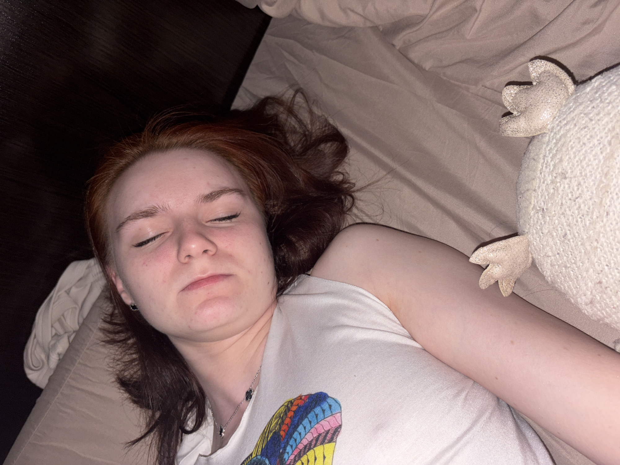 A young adult female is lying on a bed, possibly sleeping. She has reddish-brown hair, fair skin, and her eyes are closed. She is wearing a white t-shirt with a colorful graphic on the front and a silver necklace with a clover-shaped pendant. Next to her is a light-colored, textured stuffed animal with protruding ears. The bed has tan-colored bedding, and a dark brown wooden headboard is visible in the background. The location is unknown.
