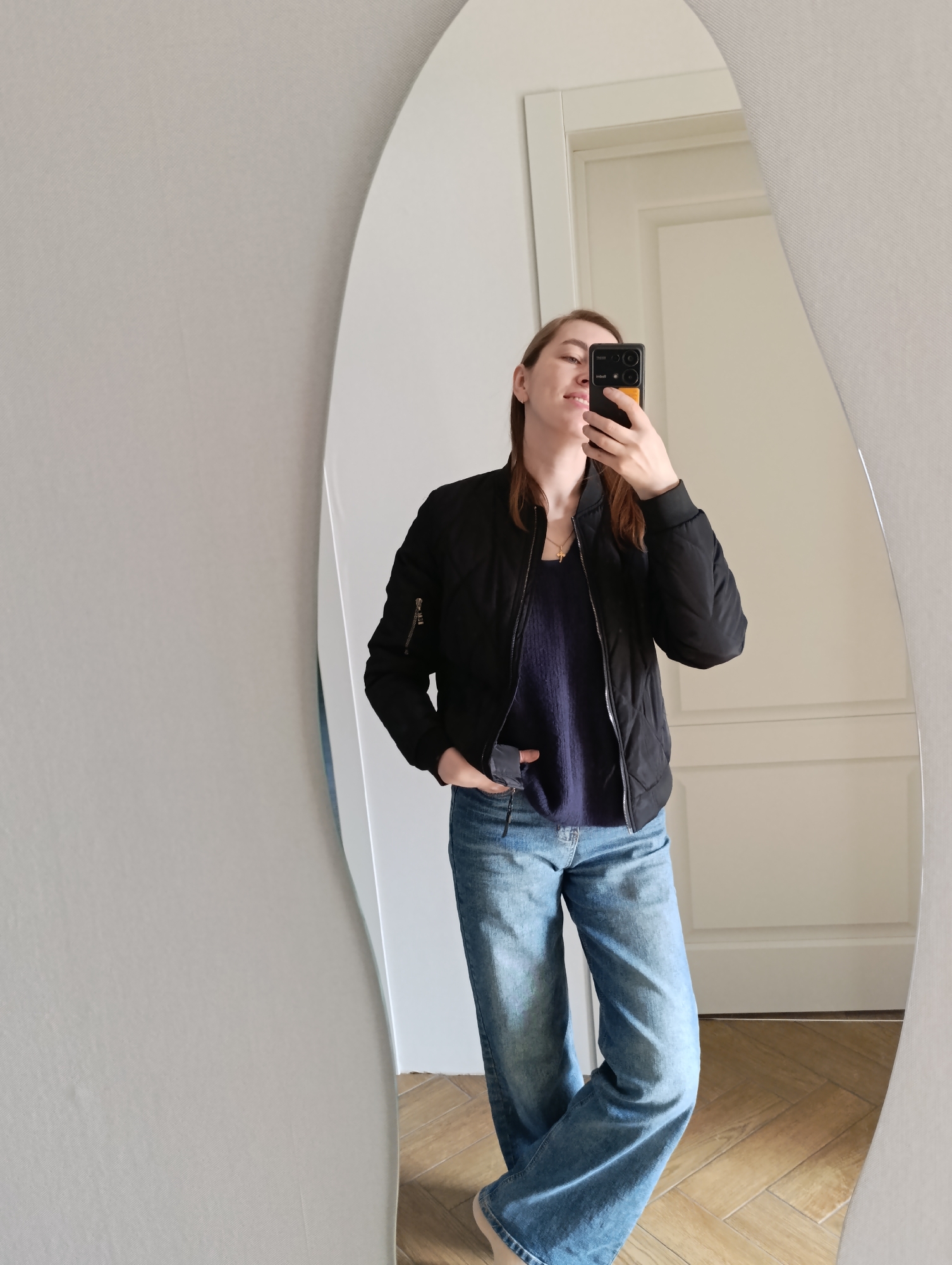 A woman is taking a selfie in front of a mirror. She is wearing a black bomber jacket over a dark blue sweater, and light blue jeans. She is holding a black smartphone in her right hand, partially obscuring her face. Her left hand is in her pocket. The mirror has an irregular, elongated shape. The background shows a white wall, a white door, and a light-colored wooden floor. The city and country are not discernible from the image.