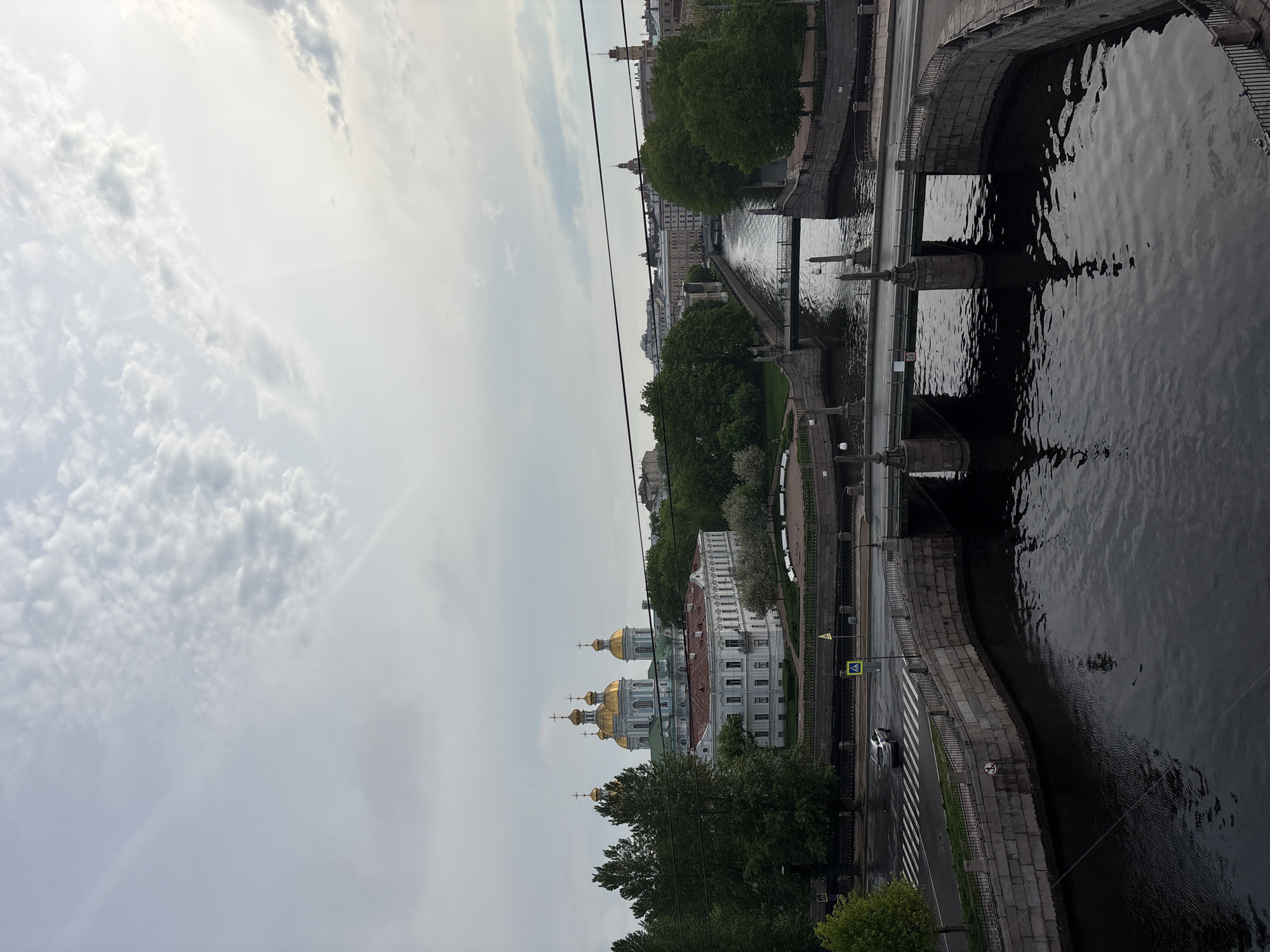 The image depicts a cityscape in Saint Petersburg, Russia, showing the Griboyedov Canal and the Saint Nicholas Naval Cathedral. The cathedral, with its distinctive blue and white façade and gold domes, is prominent on the left side of the image. The canal runs along the right, with stone embankments and bridges crossing the waterway. Trees line the canal and provide greenery, contrasting with the grey stone and overcast sky. A pedestrian crosswalk is visible alongside the canal, and a car is traveling on the road. The sky is cloudy, suggesting a diffused light.