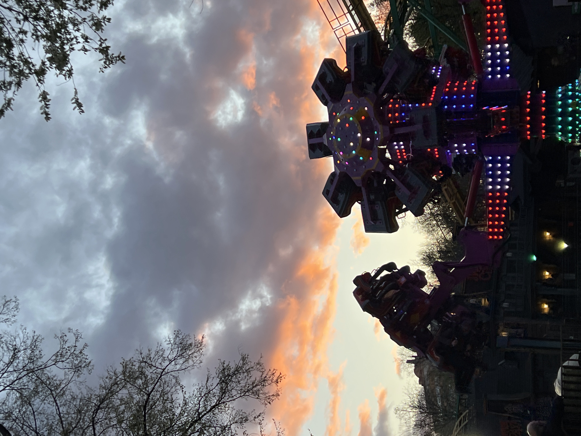 The image depicts a carnival ride against a dramatic sunset sky. The ride, featuring multiple rotating arms and platforms, is illuminated with strings of colorful LED lights, creating a vibrant contrast with the dusk lighting. Bare tree branches are visible in the lower left and right corners, suggesting an autumn or winter scene. The presence of a roller coaster track partially visible above the carnival ride indicates that the image was captured in an amusement park setting. There are some people on the ride and around it. Due to the available information, it is not possible to definitively confirm the location as the city of Perm, Russia.