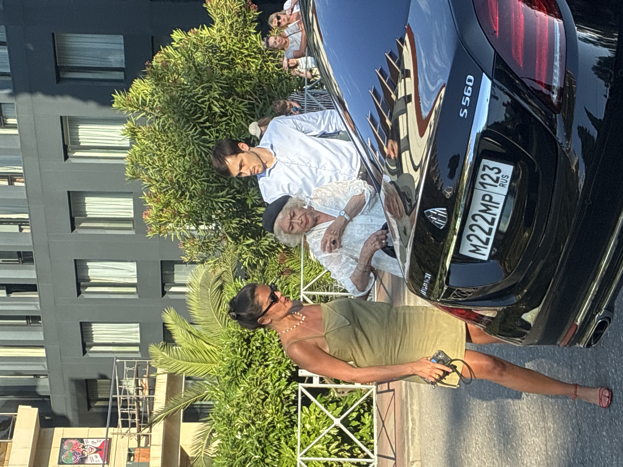 In Sochi, Russia, three individuals stand near a black Mercedes-Maybach S560 sedan parked on a street. A woman in a khaki dress and sunglasses holds a phone and a purse. Beside her, an elderly woman in a white lace top and a black beret is leaning towards the car. A young man in a light blue collared shirt stands behind the elderly woman, looking to the side. In the background, a modern building with dark gray siding and white-curtained windows is visible, along with lush green foliage. The license plate of the car reads "M222MP 123 RUS".