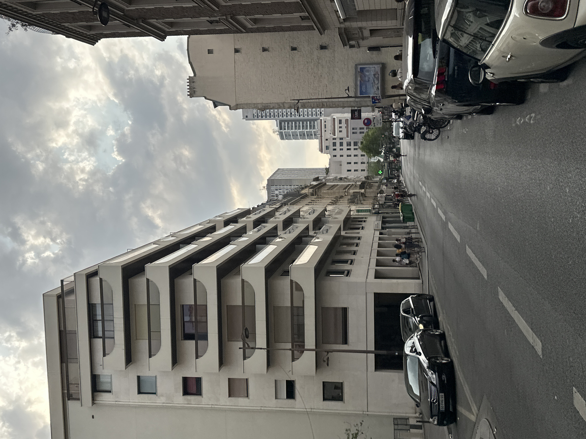 The image depicts a street scene in Paris, France, under a cloudy sky. On the left is a modern, white apartment building with numerous balconies featuring curved railings. The building has multiple floors with dark-framed windows. In front of the building, a street is lined with parked cars, including a black Mercedes-Benz sedan with a license plate reading "68-FHQ-92", and another black SUV. Further down the street, more cars and parked motorcycles are visible. People are seen walking on the sidewalk and cycling on the street.
To the right of the modern building, a series of older, narrower buildings with traditional Parisian architecture are visible. These buildings are made of brick and stone, with numerous windows and some ornate detailing. A street lamp extends from the modern building over the street. Further down the street, taller, more modern buildings are visible in the distance. The street itself is paved and marked with white lines. The overall atmosphere suggests an urban environment in a somewhat overcast condition.