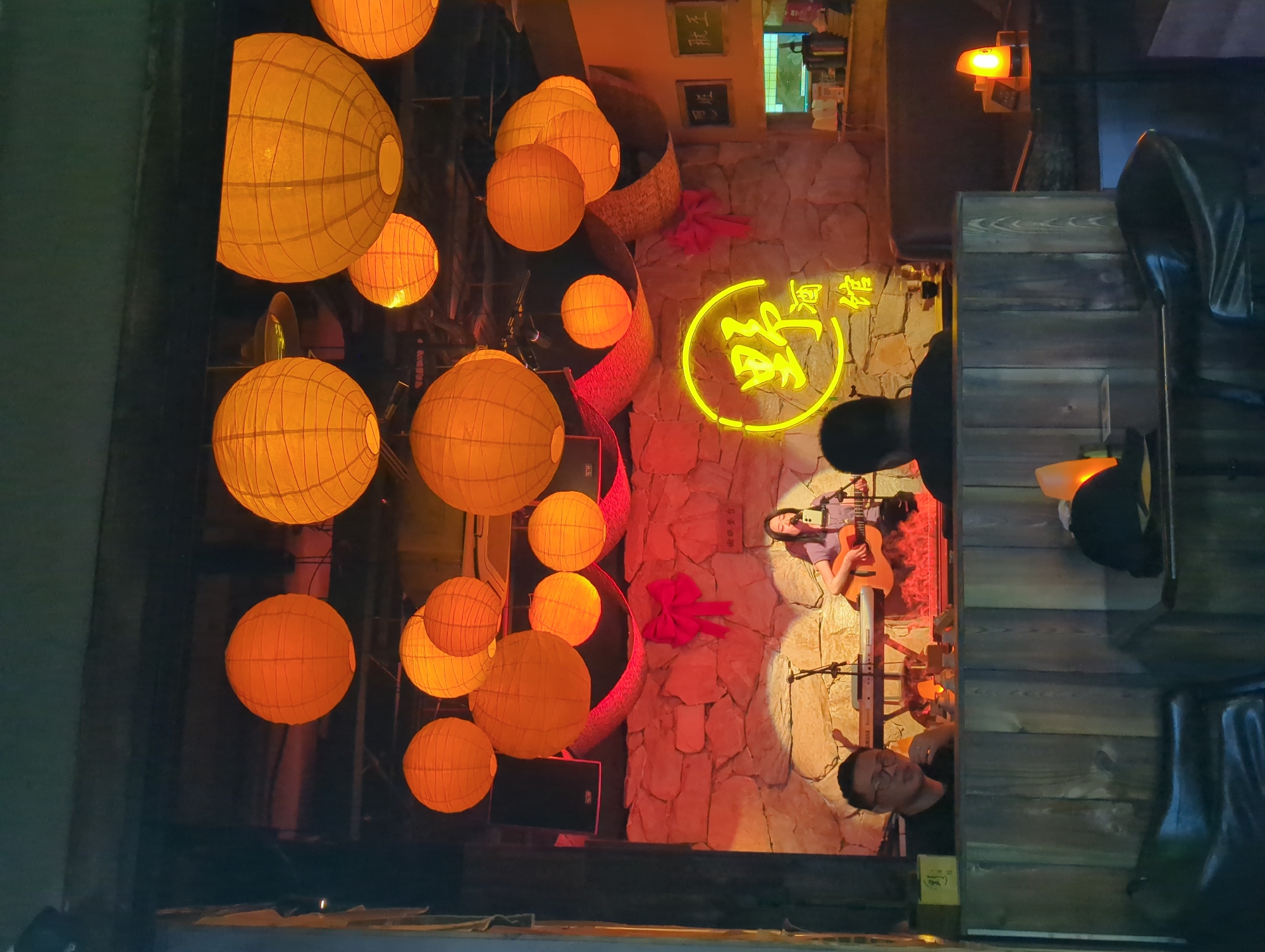 The image depicts an interior scene in Liangjiang, China. A performer plays guitar on a stage with a stone-textured backdrop, illuminated by stage lighting and a neon sign with Chinese characters. The space is decorated with numerous orange paper lanterns. In the foreground, part of an audience, including a man wearing glasses, is visible. Elements of furniture are also apparent.