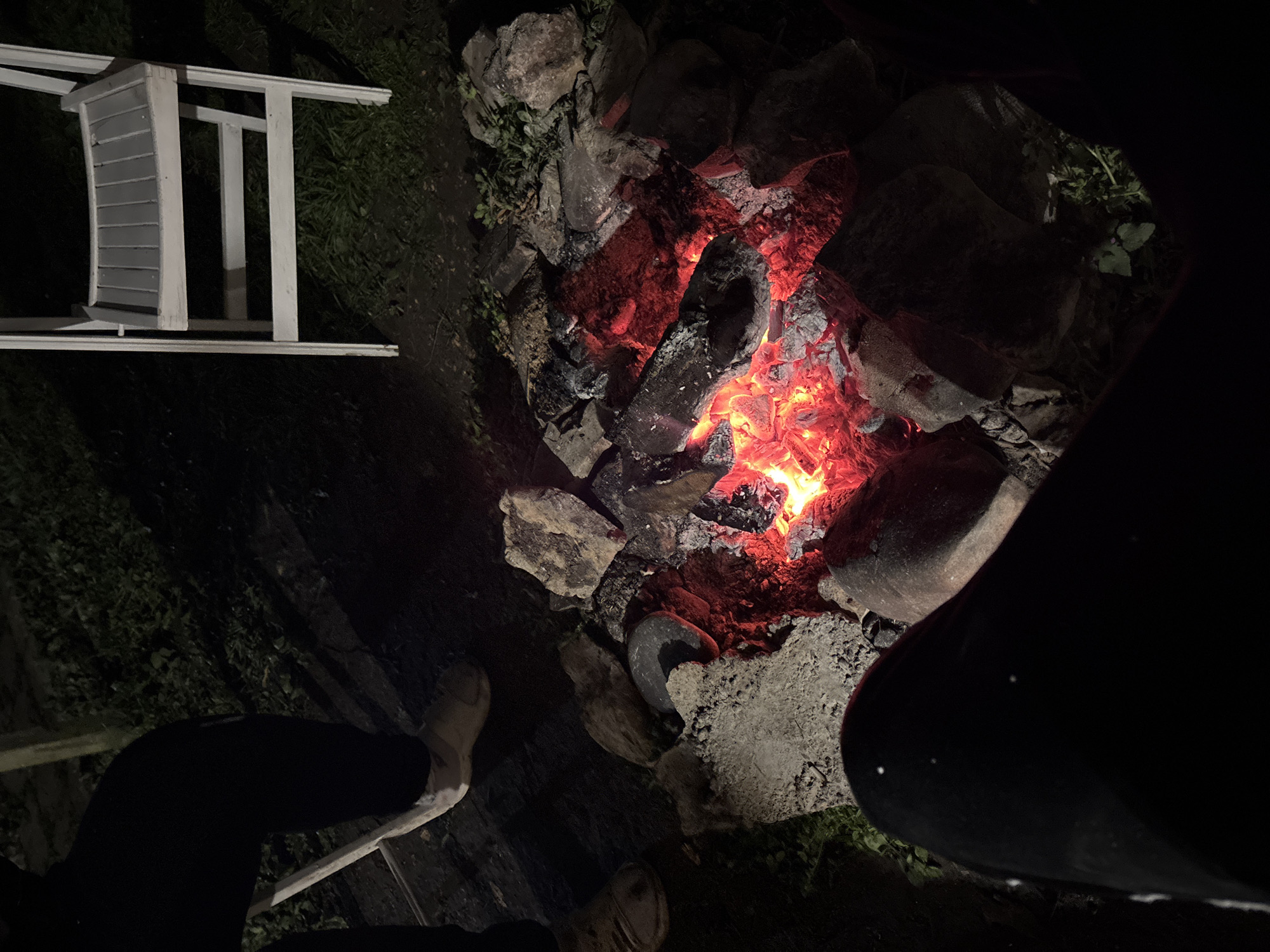 A night-time, high-angle shot depicts a stone fire pit containing burning wood, surrounded by various rocks and foliage. To the left, a partial white wooden structure is visible. Two pairs of legs and feet, clothed in dark trousers and light-colored shoes, are seen near the fire pit. The precise location is not discernible, and there is no information available to confirm whether the image was taken in Karlukovo, Bulgaria.