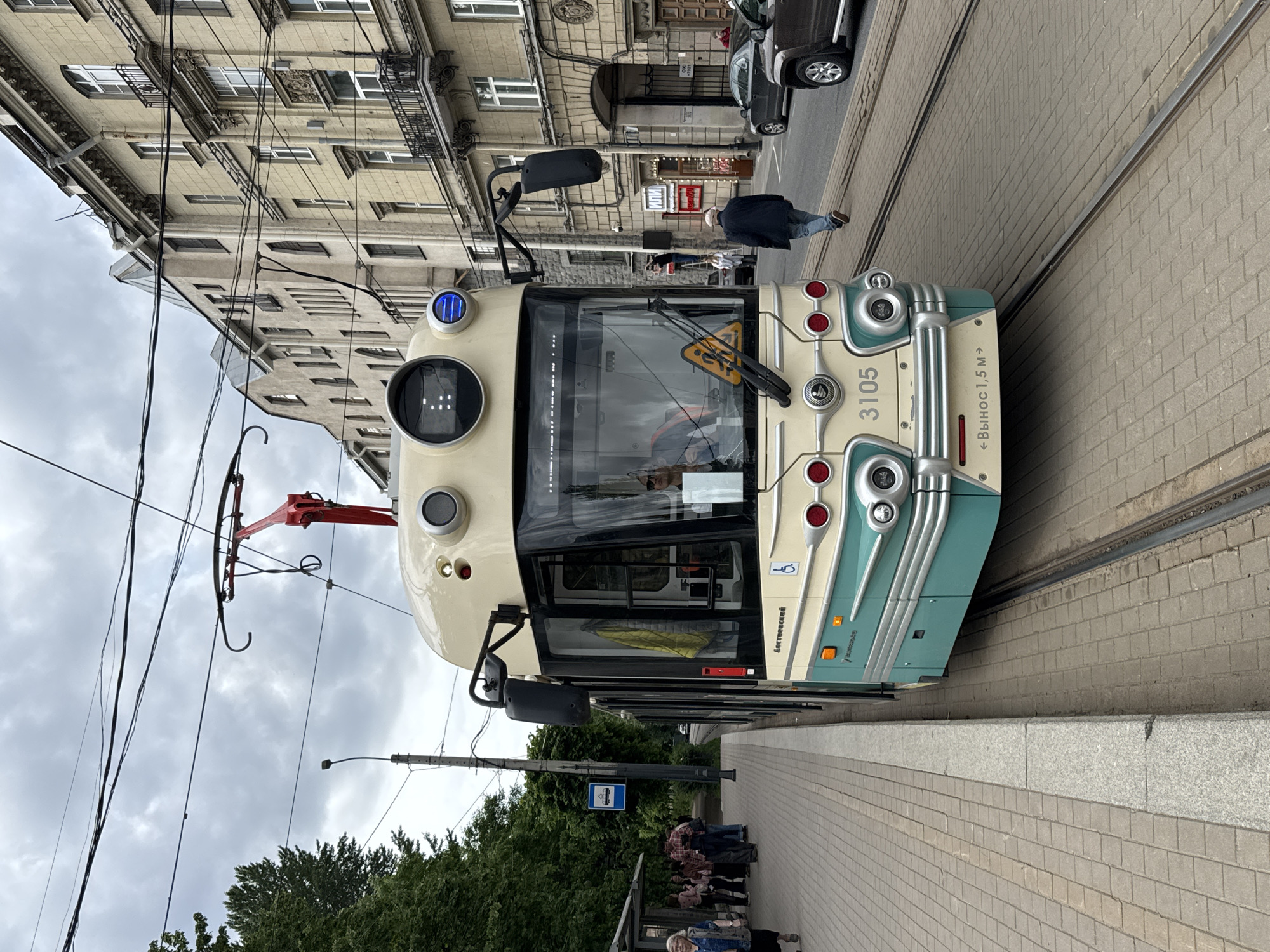 The image displays an urban street scene in Saint Petersburg, Russia, under an overcast sky. A cream and teal-colored tram, numbered 3105, is centrally featured, appearing to be stopped at a tram stop. Its red pantograph is extended upwards, connecting to a complex network of overhead electrical wires essential for its operation.
On the front of the tram, the number "3105" is clearly visible, along with the text "← ВЫНОС 1,5 М →" (OVERHANG 1.5 M →), likely a warning about its width. A yellow triangular sign featuring a pedestrian symbol is affixed to the front window. Along the tram's side, the word "Ленинградский" (Leningradsky) is readable, accompanied by a disabled access symbol.
To the right of the tram, a wide sidewalk, paved with light-colored bricks, extends into the distance. A blue tram stop sign is visible, and a group of people are gathered on the sidewalk, seemingly waiting to board or disembark. Further down the street, another person is seen walking across the tram tracks.
The background is filled with impressive, multi-story historic buildings characterized by ornate facades, balconies, and numerous windows, typical of Saint Petersburg's architecture. A dark-colored car is parked on the street near these buildings. The street itself features prominent tram tracks running alongside the sidewalk. Green trees and foliage are visible along the lower left edge of the image. The scene depicts a routine daytime moment in a historical urban setting.