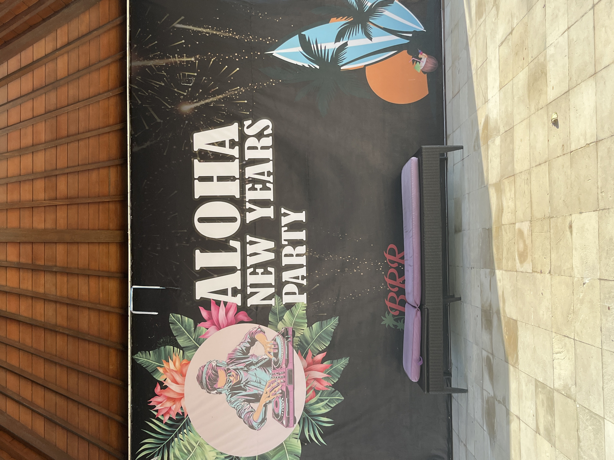 The image depicts a large, black banner with a tropical and New Year's Eve theme. The banner features the text "ALOHA NEW YEARS PARTY" in large white font. To the left, there's a circular graphic of a DJ with headphones and sunglasses, surrounded by tropical leaves and pink flowers. To the right, there is a graphic of a surfboard, palm trees, a sunset, and a coconut drink. Below the banner, a dark brown chaise lounge with two purple cushions is placed on a tiled patio. The patio is made of light-colored square tiles, some of which appear wet. Above the banner, the underside of a wooden roof structure is visible. The scene appears to be outdoors, possibly at a resort or event venue in Kuta Selatan, Indonesia, given the tropical motifs and the style of the roof.