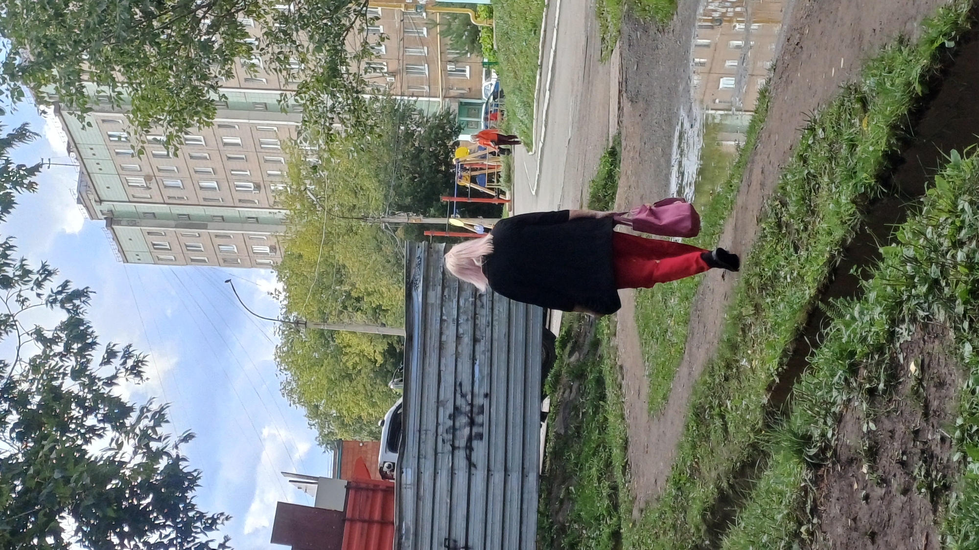 An eye-level, medium shot captures a street scene in Izhevsk, Russia, on a cloudy day. In the foreground, a woman with light pink hair walks away from the camera on a dirt path, wearing a black jacket and red pants. She carries a pink bag in her right hand. The path is flanked by green grass and a drainage ditch. In the mid-ground, a large pile of corrugated metal sheets lies to the left, with a red truck visible behind it. To the right, a playground with yellow and red equipment is set up, and another woman in an orange jacket walks further down the street. A multi-story apartment building, typical of Soviet-era architecture, dominates the background, with a pale green and beige facade. The street is wet, indicating recent rain, with reflections visible in puddles. Trees and utility poles are scattered throughout the scene.