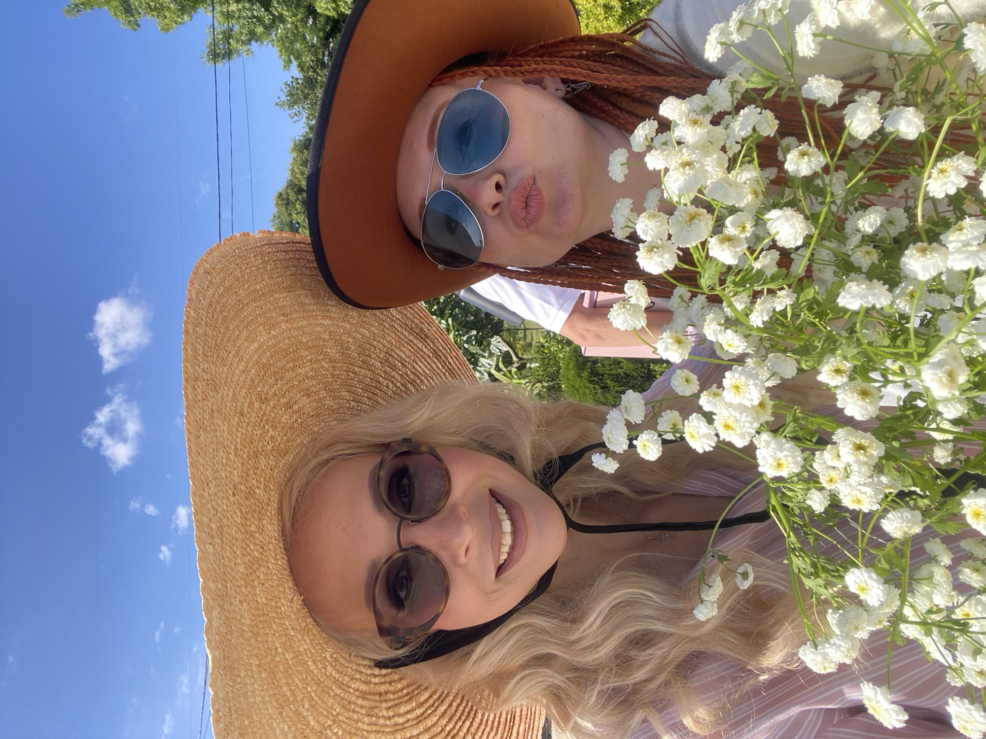 Two women are posing for a selfie in front of a bright blue sky with sparse white clouds. Both women are wearing wide-brimmed hats and sunglasses. The woman on the left has blonde, wavy hair and is wearing a pink and white striped shirt. She is smiling and holding a bouquet of white flowers. The woman on the right has reddish-brown braided hair and is wearing a brown hat and aviator sunglasses. She is puckering her lips as if for a kiss and is also holding the bouquet of white flowers. The background shows greenery, suggesting an outdoor setting, possibly a park or garden. There are no specific markers to identify the location as Sochi, Russia.