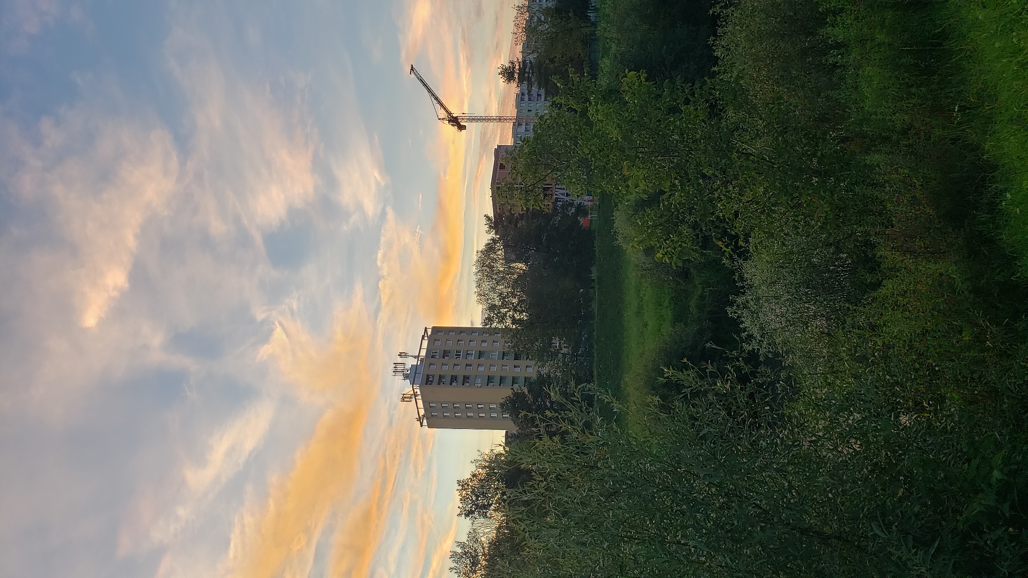 The image depicts a sunset over a residential area in Prešov, Slovakia. In the foreground, lush green foliage fills the lower portion of the frame. Behind the trees, a tall, light yellow apartment building with multiple windows and antennas on its roof stands prominently. To the right of this building, a construction crane is visible, along with other apartment buildings in the background. The sky is a mixture of blue and light orange, with wispy clouds, suggesting the late afternoon or early evening. The scene is devoid of any people or visible activity.