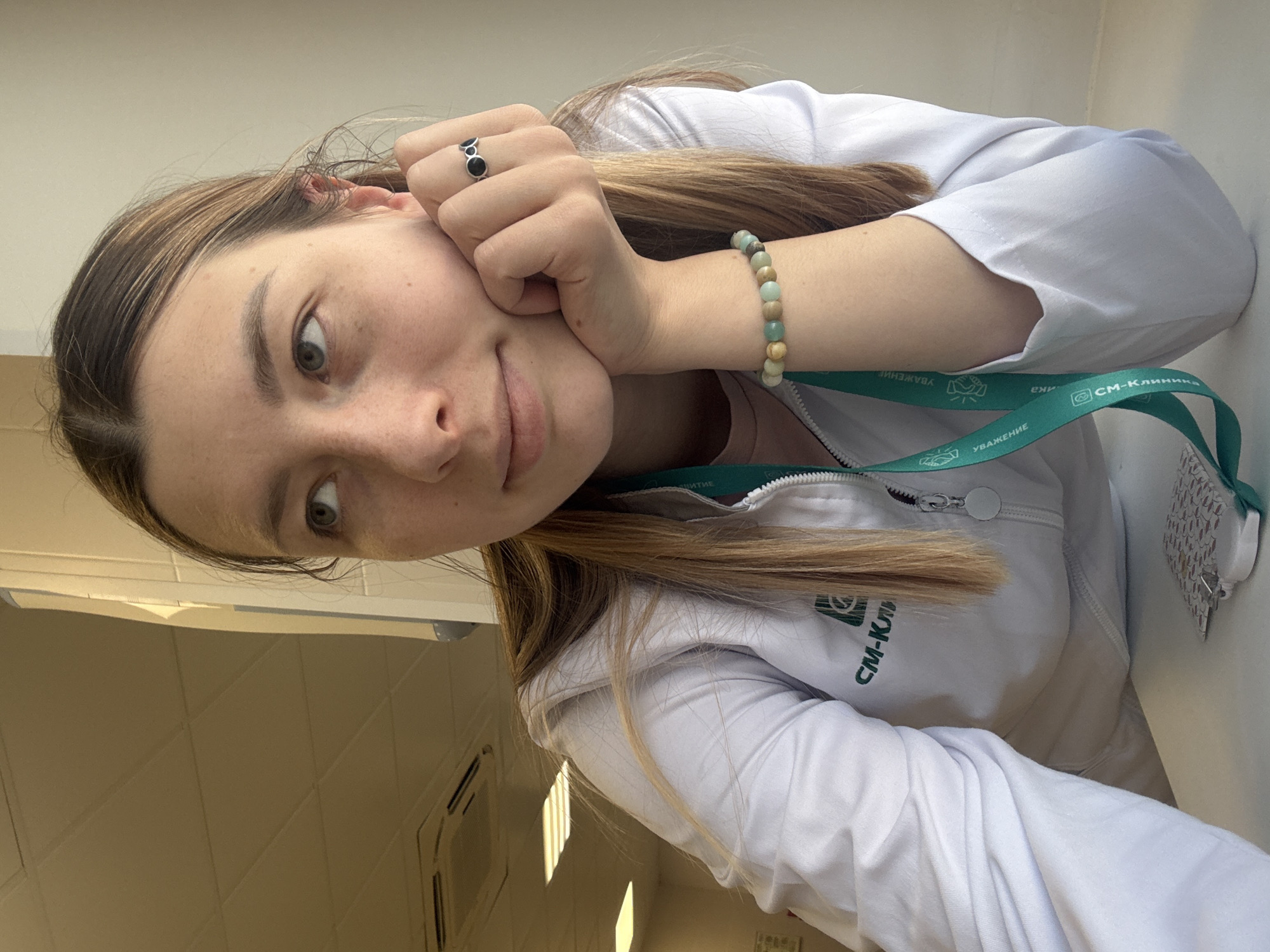 A young woman is captured in a head-and-shoulders shot, rotated approximately 90 degrees counter-clockwise. She is indoors, presenting a neutral to slightly thoughtful expression as she looks directly at the camera, resting her head on her left hand.
She is wearing a white lab coat or medical uniform jacket, which is partially zipped. A green lanyard hangs around her neck, prominently displaying the text "СМ-Клиника" (SM-Klinika), identifying a Russian medical clinic, and also the word "УВАЖЕНИЕ" (RESPECT). Her hair is long, light brown with blonde highlights, falling over her shoulders. Accessories include a black ring with two circular black stones on her left ring finger and a multi-colored beaded bracelet on her right wrist.
The setting appears to be a brightly lit indoor environment, possibly a clinic or office. The background includes a light-colored wall, a ceiling with visible panels and light fixtures, and bright light entering from an unseen window or overhead source, suggesting it is daytime. The image appears to be a selfie.