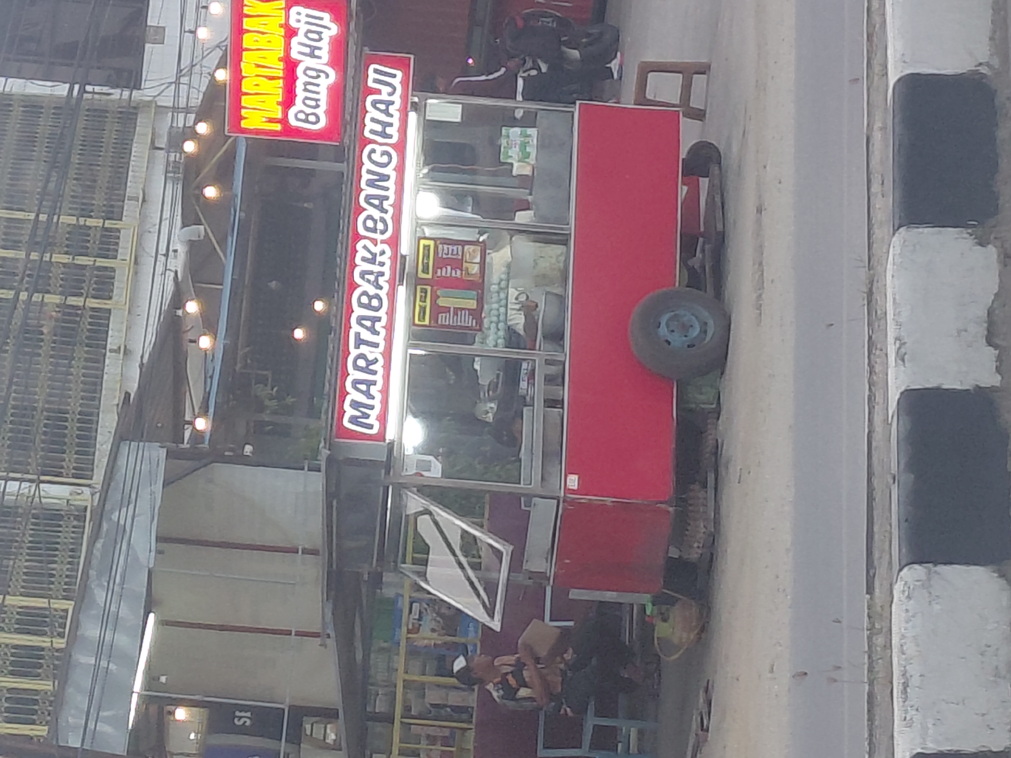 The image shows a Martabak Bang Haji food cart on a roadside in Kendari, Indonesia. The food cart is red with a white canopy and illuminated signage. Two men are visible near the cart; one is sitting on a blue bench to the left, and another is standing to the right. The background consists of a building with metal shutters and street lighting. A motorcycle is parked to the right of the cart. The scene appears to be outdoors, possibly in the evening, given the artificial lighting. The foreground shows a road and a concrete divider.