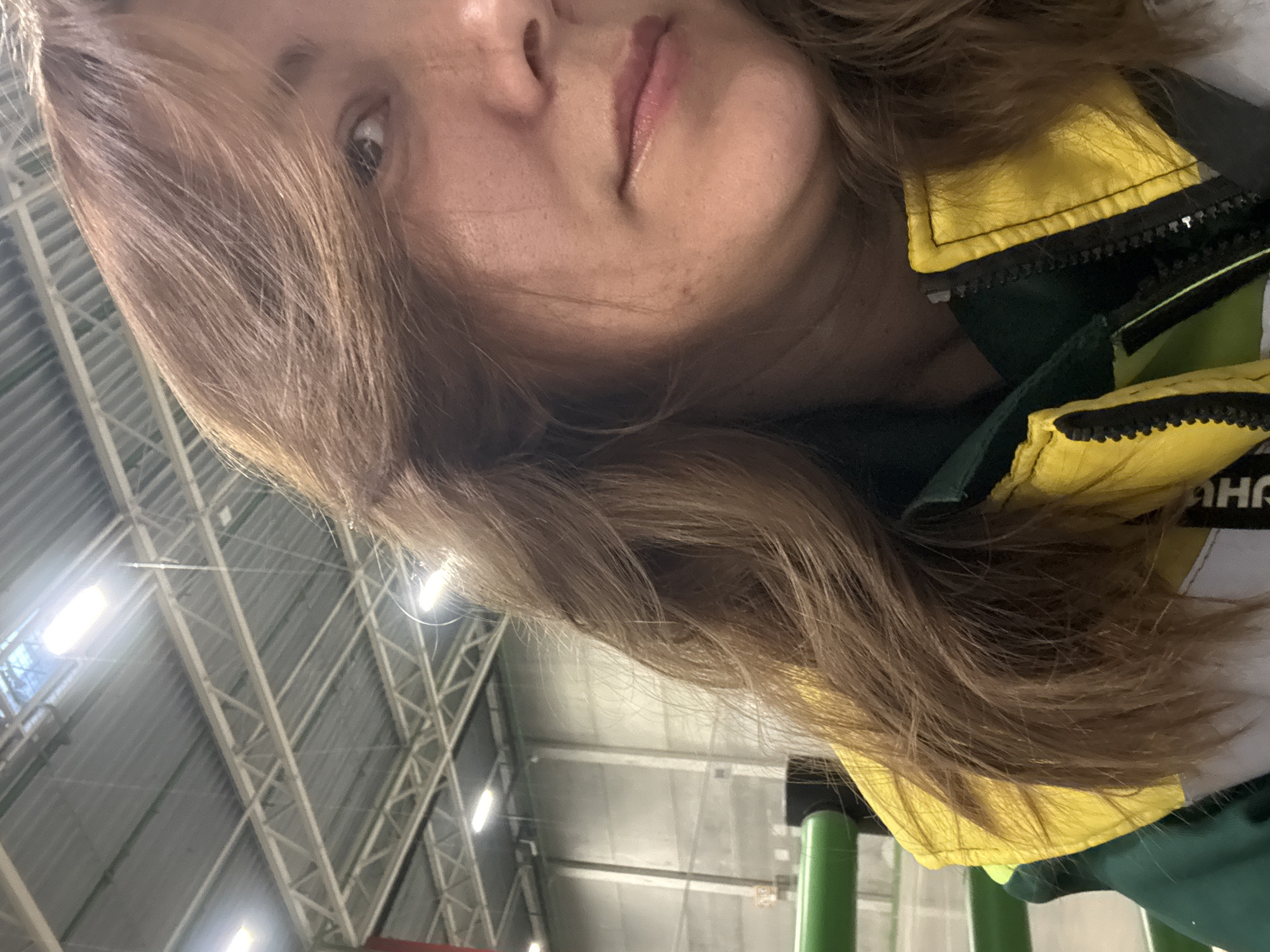 A close-up, angled shot of a person's face, likely a selfie. The person has fair skin, light brown hair, and is wearing a dark green and yellow jacket. The background suggests an indoor space with a high, industrial-style ceiling featuring metal beams and artificial lighting. A green pole or structure is visible in the lower part of the frame. There is no indication of Lupanovo, Russia.