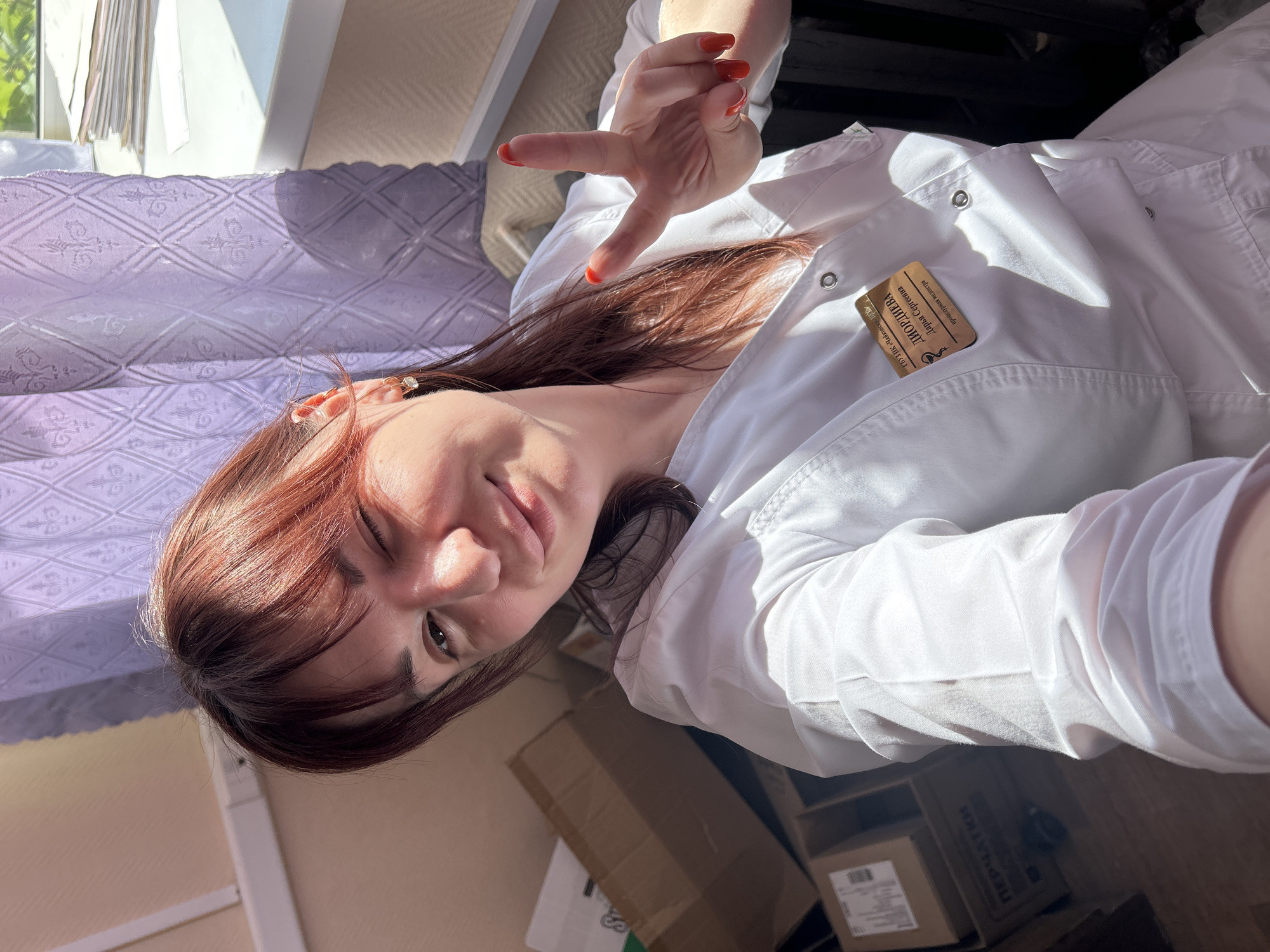 A woman with auburn hair, wearing a white lab coat and a name tag labeled "ЛИМОРИНА Дарья Сергеевна," is taking a selfie. She is winking with her left eye and holding up a "peace sign" with her right hand. A window is visible in the background, covered by a light-purple patterned curtain. The room has cardboard boxes on the floor. The location is in Фоки, Russia.