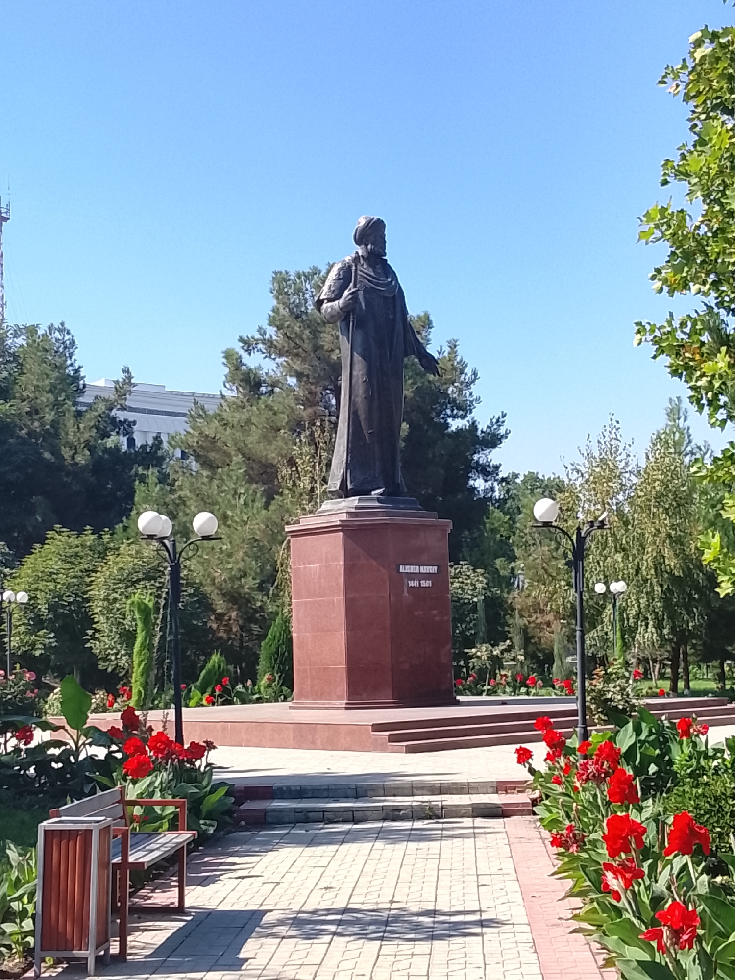 The image shows a statue of a man in traditional attire standing on a red granite pedestal in a park. The statue is located in Fergana City, Uzbekistan, likely at a memorial or historical site. In the foreground, there is a paved walkway lined with red flowers and green foliage. A wooden bench and a trash can are also visible in the foreground. The background features trees, a building, and a clear blue sky. The overall scene depicts a well-maintained public space with a focus on historical commemoration and natural beauty. The inscription on the pedestal appears to be in a non-Latin script, but the dates 1441 and 1509 are visible, suggesting a historical figure.