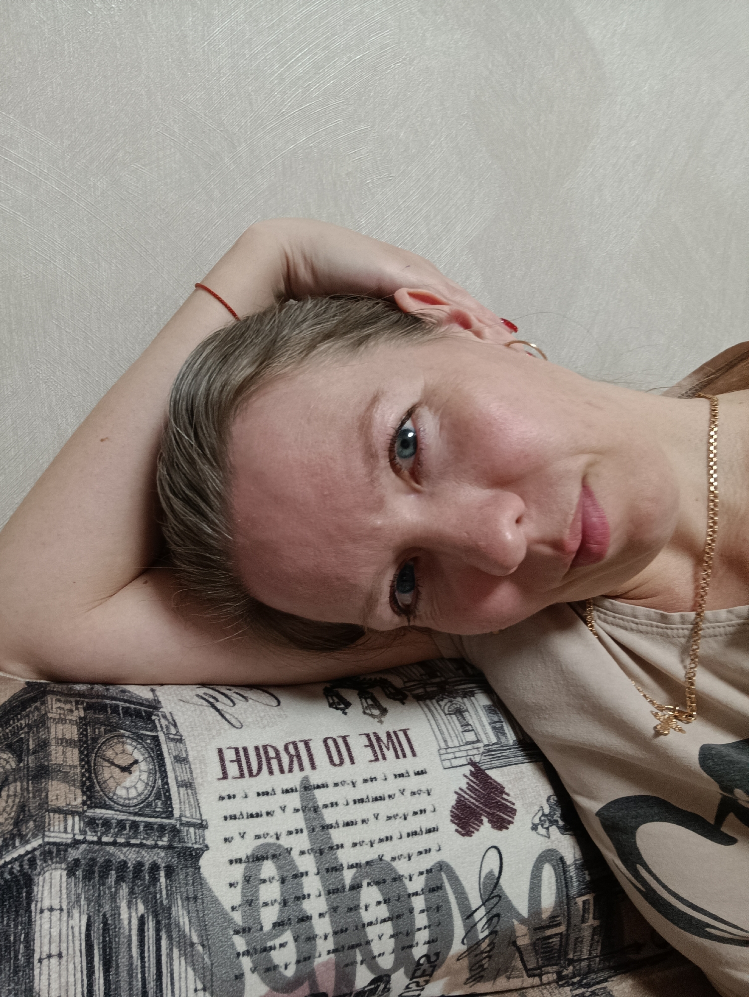 Here is a detailed description of the image:
**People:** The image shows a woman with fair skin, light blue eyes, and graying hair. She is wearing a gold necklace.
**Environment:** The background is a plain, light-colored wall. She is resting her head on a pillow or cushion.
**Objects:** The pillow/cushion has a patterned design featuring an image of the Big Ben clock tower. The pillow also has text that reads "TIME TO TRAVEL" in reverse.
**Actions/Interactions:** The woman is lying down and looking at the camera with a slight smile. She rests her head on her arm.
**Details:** She has a red bracelet on her wrist, and red nail polish on the fingernails. She has gold earring.
**Location:** The location is unknown.