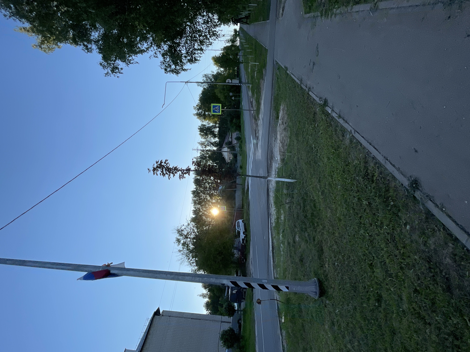 The image depicts a street scene in the settlement of Krasnye Baki, Russia, bathed in the warm light of a setting or rising sun. The perspective is from a sidewalk looking towards a road intersection. On the left side of the frame, a light-colored apartment building is partially visible. In the foreground, a lamppost stands with a Russian flag attached to it.
The road is paved, with a crosswalk visible at the intersection. Several vehicles are parked or driving in the background. The sun is low in the sky, creating a bright flare as it shines through the trees. A pedestrian crossing sign is prominently displayed on a green post near the intersection. The sidewalk is concrete, with a grassy area bordering it. Trees line the right side of the sidewalk, creating a shaded path. The sky is a clear, light blue.