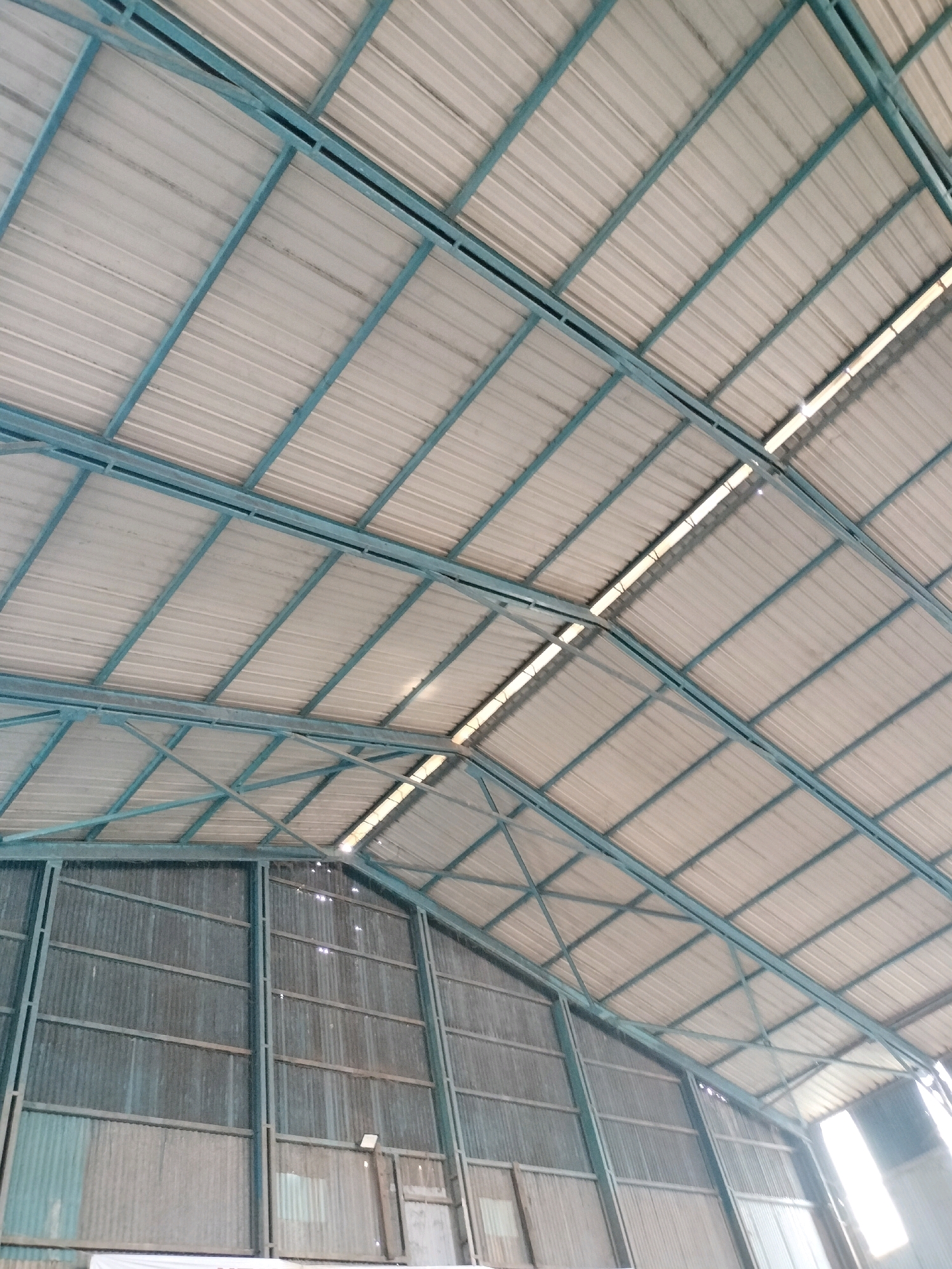 The image shows the interior of a large industrial or warehouse-like structure, likely in Batam, Indonesia. The perspective is looking upwards at the corrugated metal roof, supported by a teal-colored metal framework. Natural light filters through translucent sections of the roof, creating illuminated strips. The lower portion of the image reveals the corrugated metal walls of the structure, with some visible vertical support beams. The scene is devoid of people or any discernible activity. The environment appears utilitarian and industrial.