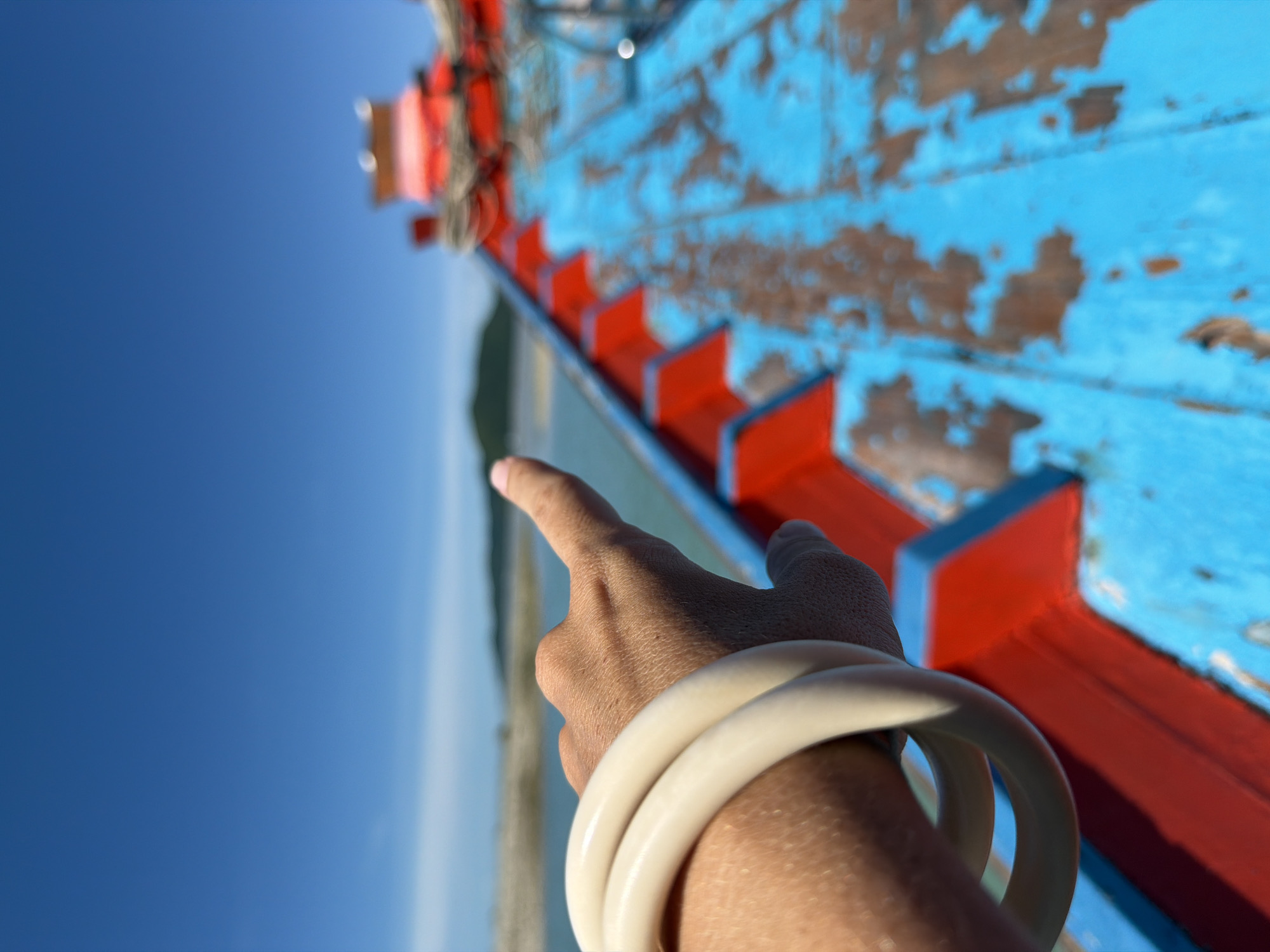 A hand wearing two cream-colored bangles points towards a distant island across the water. The hand is positioned in the foreground, on the left side of the image. The background shows a bright blue sky above a body of water and a green island. The foreground is dominated by the deck of a boat painted in vibrant blue and red, showing signs of wear. The boat appears to be in motion, with the water and distant scenery slightly blurred. The scene is set in Baan Thong Krut, Thailand.