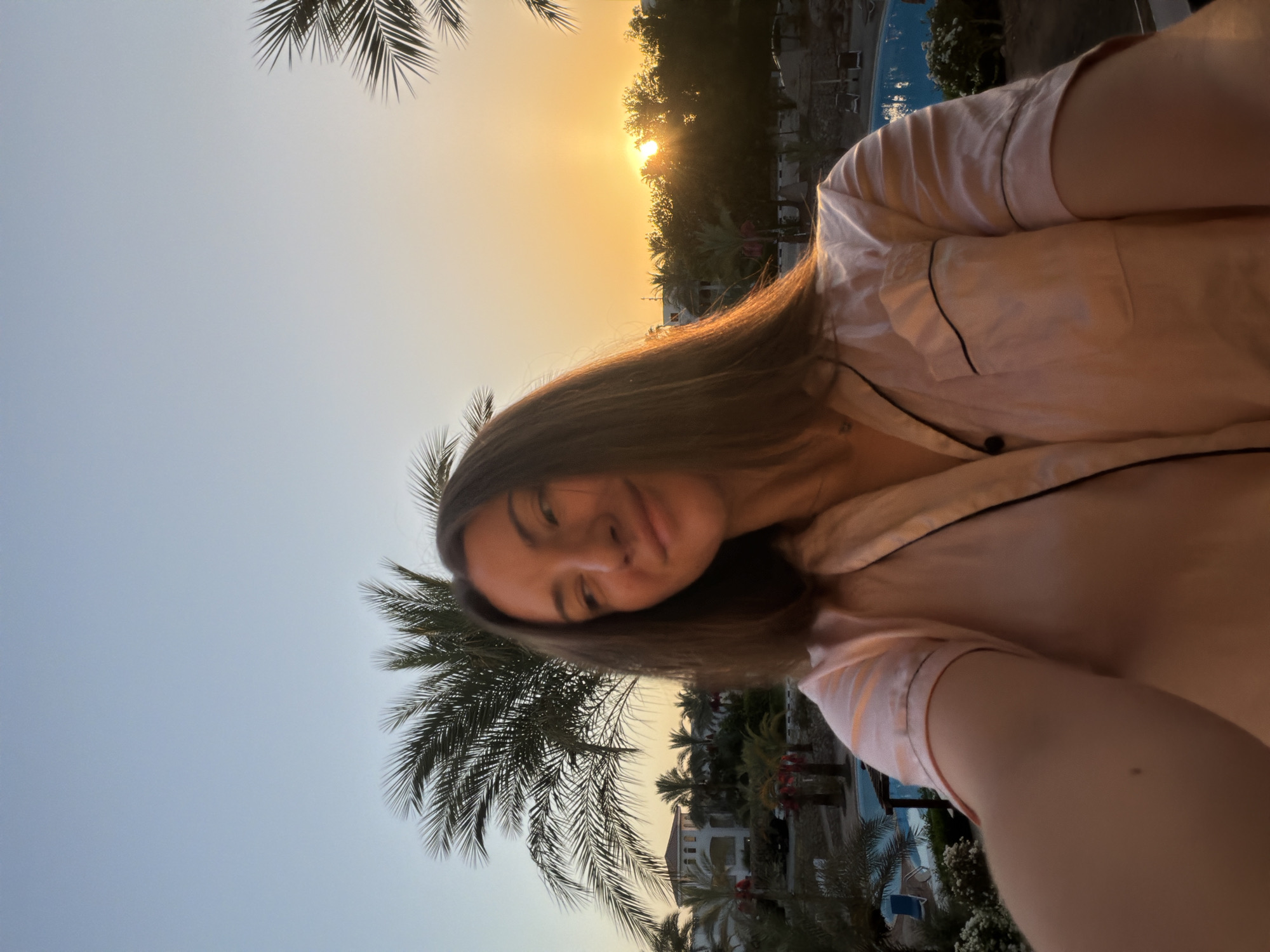 A woman is captured in a selfie on a balcony overlooking a resort pool at sunset in Sharm El Sheikh, Egypt. The woman, with brown hair and wearing a light pink pajama top, smiles towards the camera as the setting sun casts a warm glow. Palm trees and resort buildings form the backdrop. The sky transitions from a clear blue to a vibrant orange hue as the sun dips below the horizon, partially obscured by trees. The pool area, with lounge chairs and umbrellas, is visible in the lower portion of the frame.
