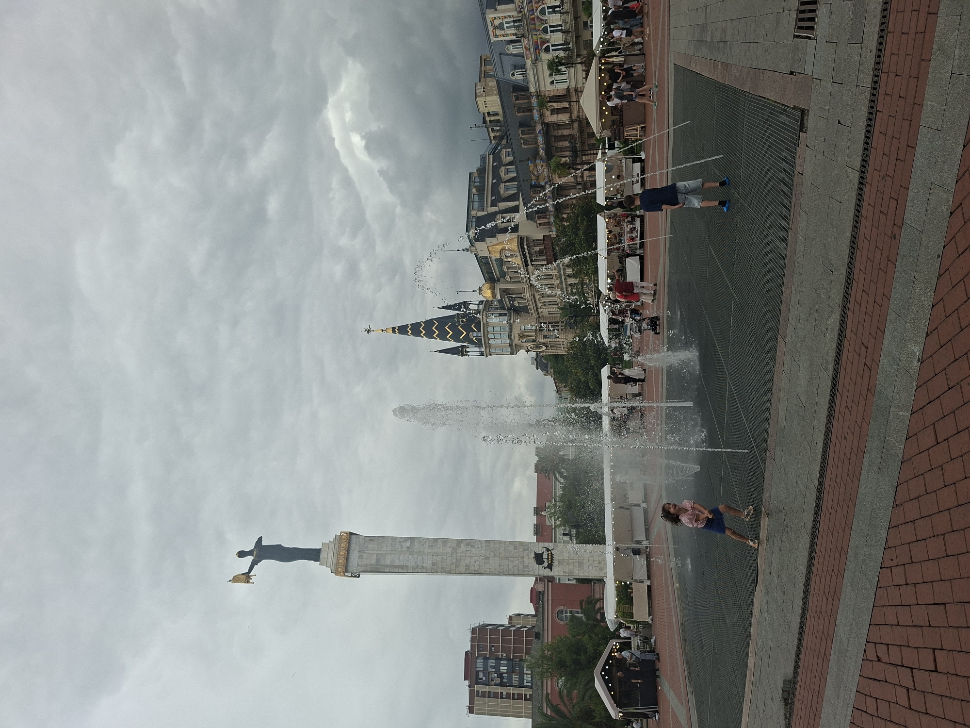 The image depicts a public square in Batumi, Georgia, under an overcast sky. In the background, a tall, white monument topped with a statue of a figure holding a golden object stands prominently. Adjacent to the monument, a distinctive building with a dark, pointed roof adorned with golden accents is visible, alongside other multi-story buildings with traditional architecture.
In the foreground, a water fountain sprays water, creating a playful environment. Two children are interacting with the fountain; a young girl in a pink shirt and blue skirt is running and laughing, while a boy in a blue shirt and grey shorts stands near the water jets. Surrounding the fountain area, there are white market stalls set up, with people browsing and interacting. The ground is paved with red and grey bricks. The overall scene suggests a lively public space with cultural landmarks and recreational activities.