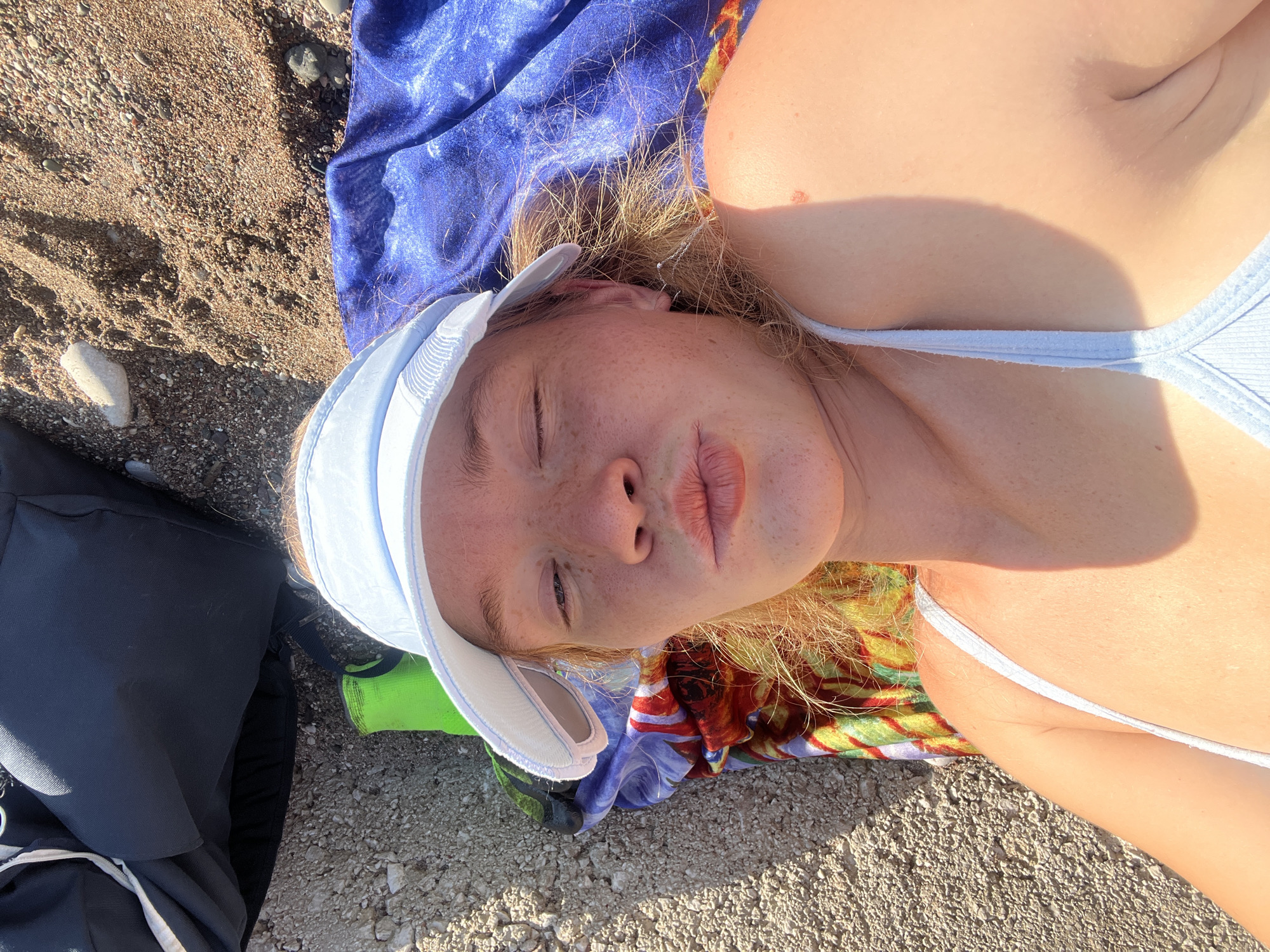 A young person with fair skin and freckles, wearing a light blue bikini top and a white baseball cap, lies on a beach blanket. Their eyes are closed, and they appear to be resting. Their blonde hair is spread out on the blue blanket, which is situated on a pebbly beach. To the right of the person, a dark backpack rests on the sand. The image is oriented such that the person's head is towards the bottom right, and their feet are towards the top left. The lighting suggests it is daytime, possibly with the sun casting shadows. The overall scene depicts a relaxed moment on a beach.