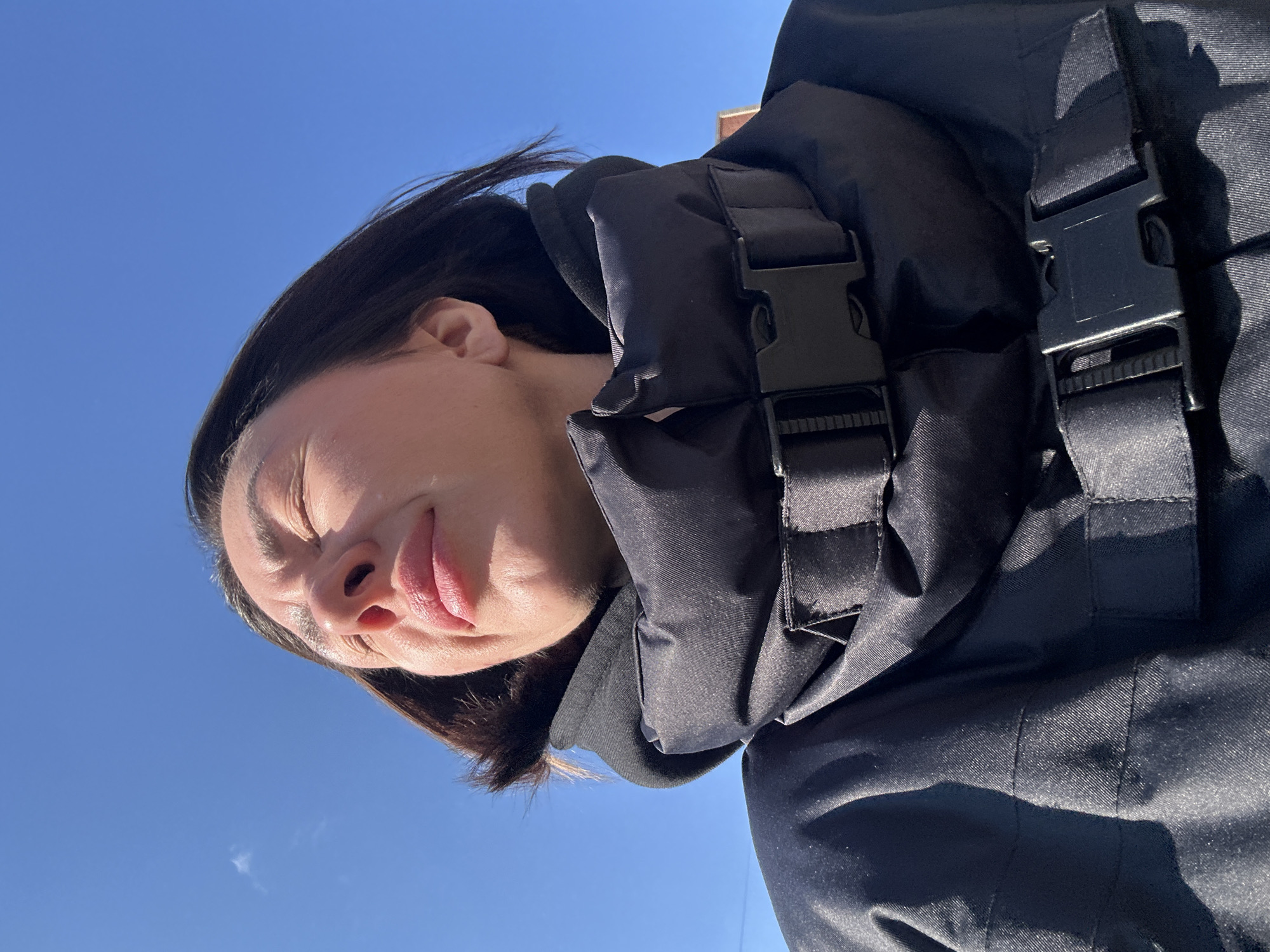 A low-angle, slightly rotated shot depicts a person with closed eyes facing the sun. They are wearing a black jacket with visible buckle closures. The background is a clear, cloudless blue sky. The location is not identifiable as Votkinsk, Russia, from the available visual information.