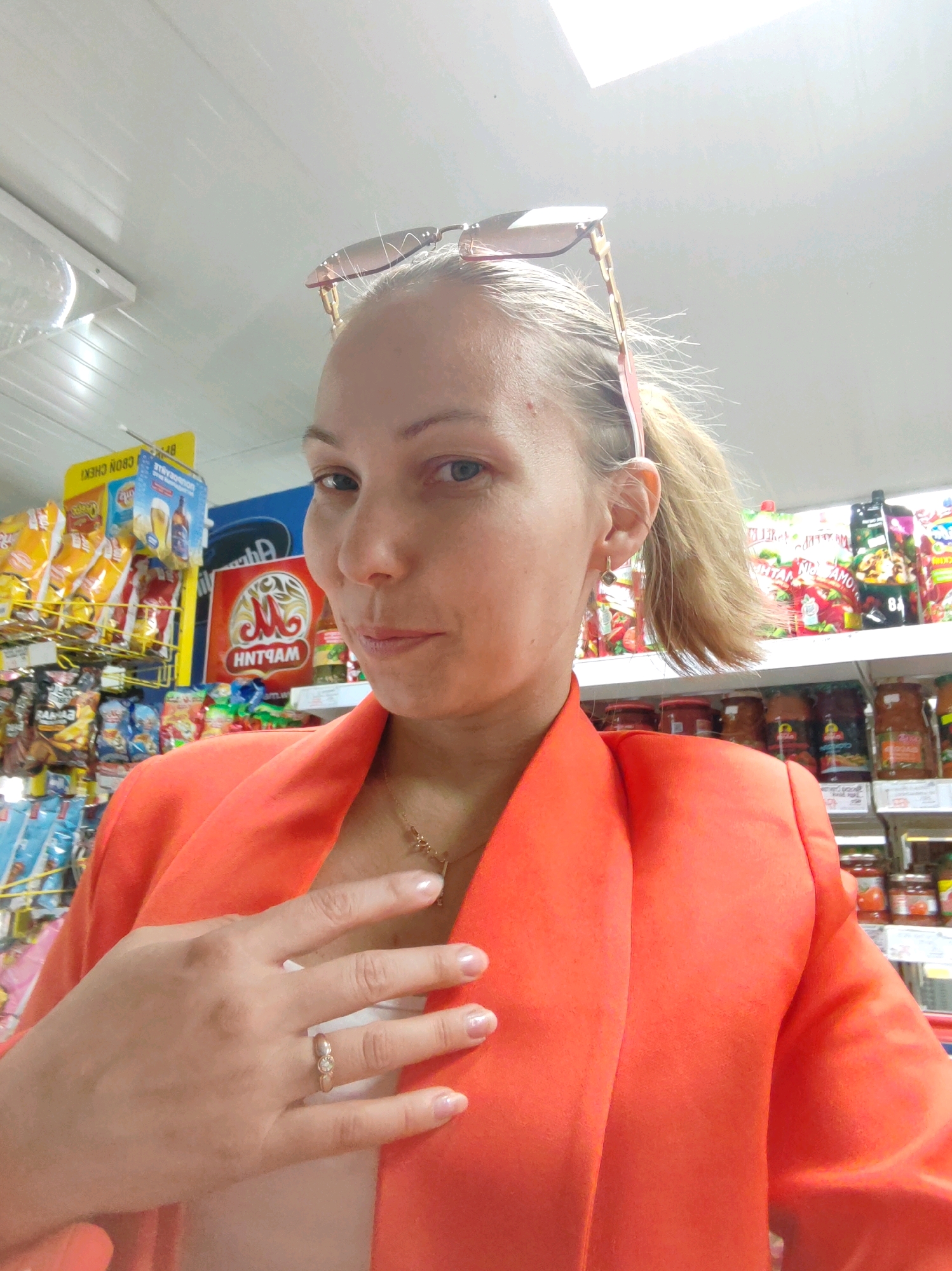 A woman with blonde hair pulled into a ponytail and sunglasses on top of her head is taking a selfie in what appears to be a grocery store aisle. She is wearing an orange blazer and a necklace. Her left hand is raised, showing a ring with three small stones. Shelves with various food products, including chips and jars, are visible in the background. The lighting suggests an indoor setting. There is no indication of the specific location within the city of Чухлэм, Russia.