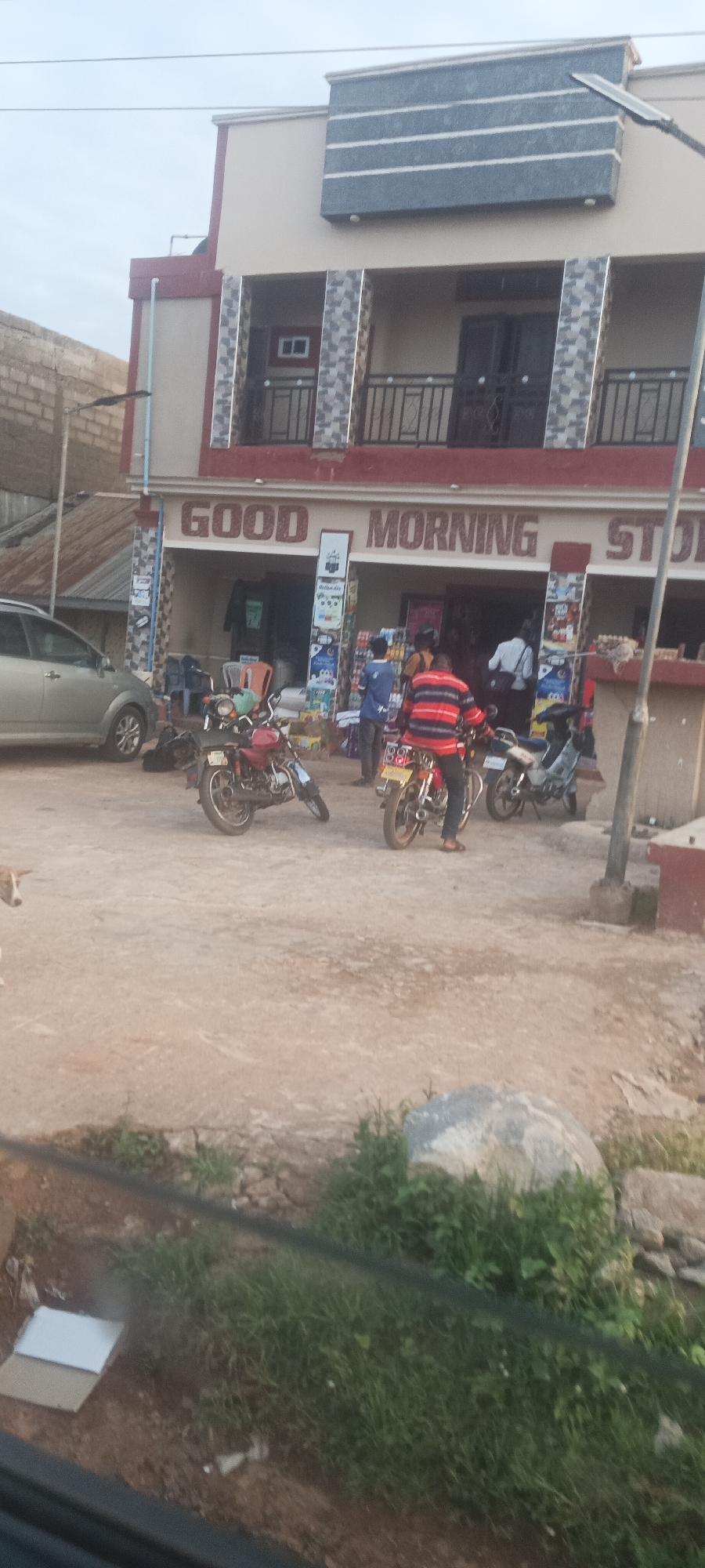 The image depicts a street scene in Mangu Town, Nigeria. The "Good Morning Store" is visible with several motorcycles parked in front. There are people milling about in front of the store, with one individual wearing a striped shirt seated on a motorcycle. A car is parked on the left. The area is unpaved and dusty. Vegetation is visible in the foreground. The building is a two-story structure. A dog appears in the left foreground.