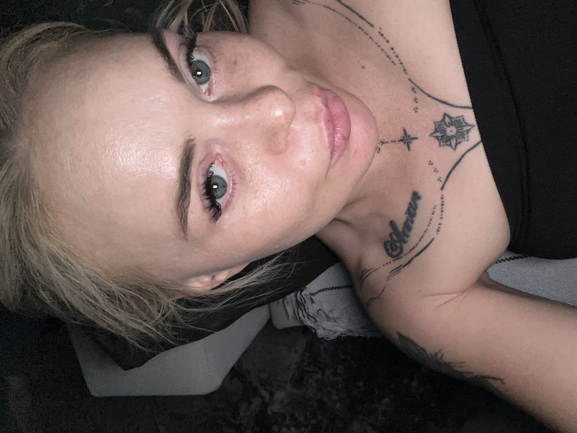 The image depicts a fair-skinned individual, likely female, captured in a close-up, high-angle shot. The subject has blonde hair, styled messily, and blue-grey eyes looking directly at the camera. Prominent dark eyelashes and shaped eyebrows frame the eyes. The skin shows visible freckles across the nose and cheeks, and a subtle sheen is present on the forehead, nose, and chin. The lips are full, light pink, and slightly parted.
The individual wears a black, strapless garment visible across the chest and upper arms. Multiple black tattoos are present on the upper torso. On the right clavicle area, the word "Amore" is written in script, accompanied by dotted lines and a small four-pointed star. Below this, centered on the sternum, is a circular geometric design resembling a stylized compass rose or mandala. Faint lines extend from these designs. A partially visible, dark, feather-like tattoo is on the left upper arm/shoulder.
The background is dark and out of focus. To the upper left and behind the subject's head, a light grey or off-white textured fabric object, possibly a pillow or blanket with visible fringe, is partially discernible. The lighting is soft and appears to originate from the front or above.
The location is Unknown, Unknown.