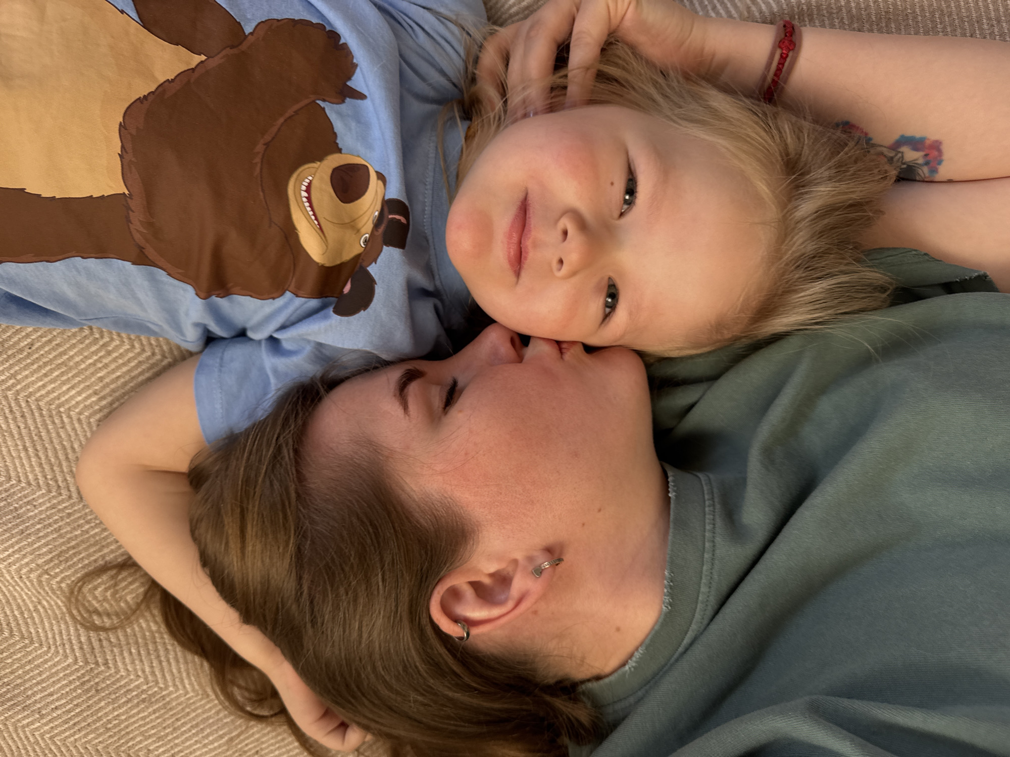 A top-down, eye-level shot depicts a woman and a young child lying down on what appears to be a rug or carpet. The woman has her face turned to the side, with her forehead near the child's face. She has brown hair, fair skin, and multiple piercings in her ear. The child has blonde hair, fair skin, and is looking directly at the camera. The child is wearing a light blue t-shirt with a large brown bear graphic on it. The woman is wearing a muted green shirt. The exact location of the city of Ферма, Russia is not determinable based on the provided image.
