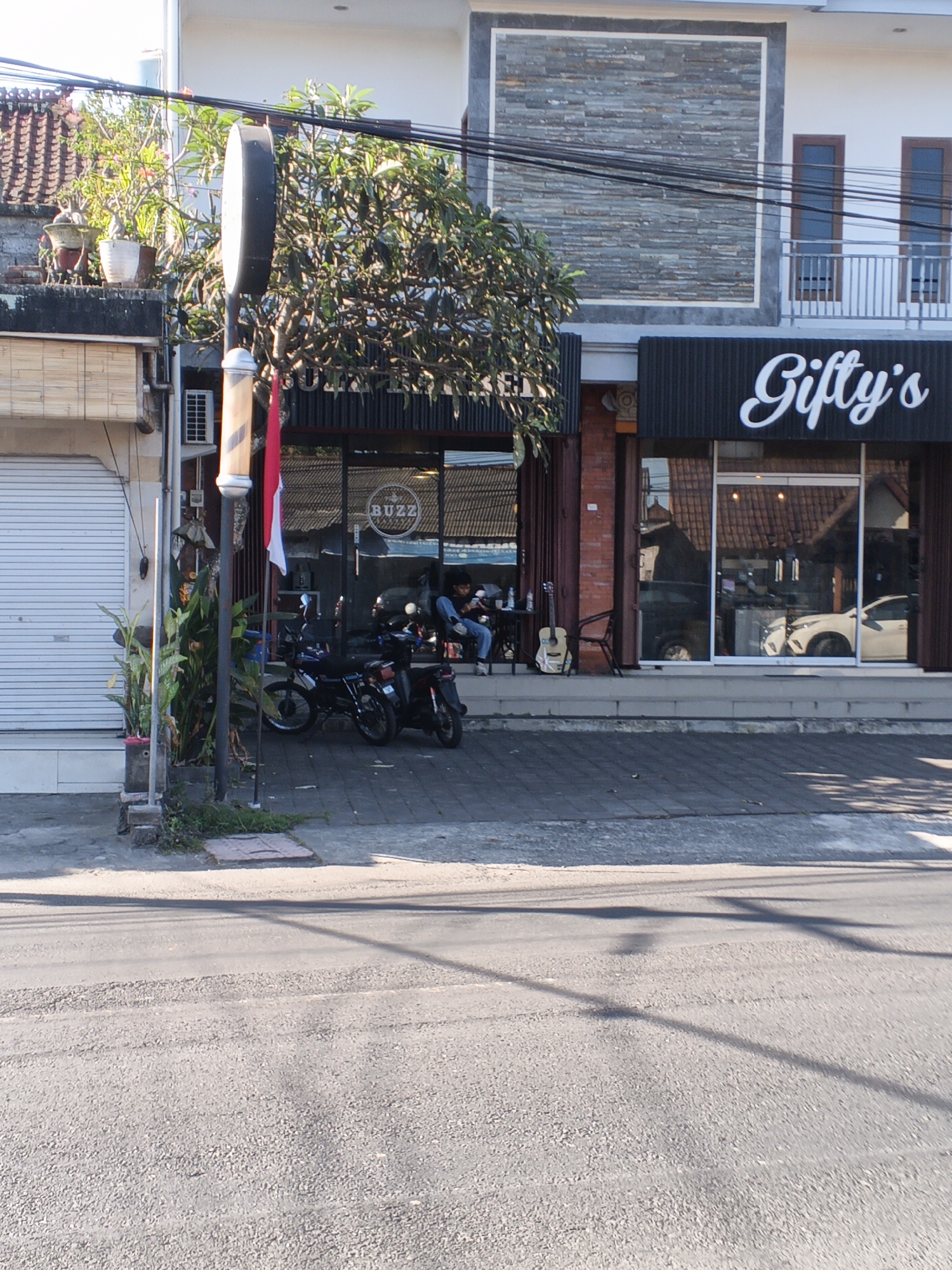 This is a street-level, eye-level, medium shot capturing the facade of two businesses in Sukawati, Indonesia. The business on the left has a white roll-up door and a red and white flag hanging from a pole with a barber pole detail. Plants in planters are visible to the left of the entrance. The business on the right, identified by the white cursive text "Gifty's" on a black awning, has a large glass storefront. Inside, a white car is visible. In front of "Gifty's," a person is seated at a small table with a guitar standing next to it. Parked motorcycles are situated in front of both businesses. The street in the foreground is paved with asphalt, and a tiled sidewalk runs in front of the businesses. Electrical wires are strung across the top of the frame.