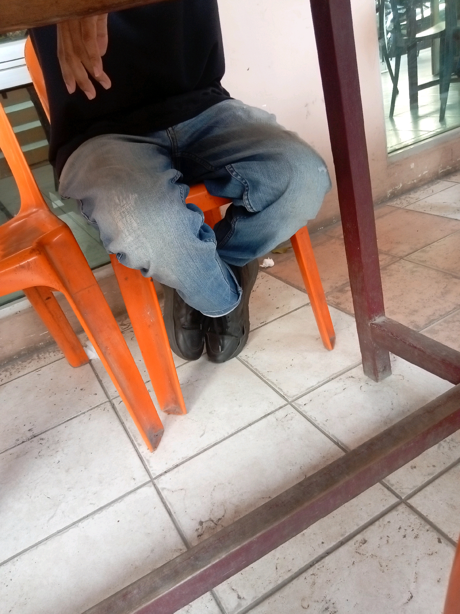 The image shows a person sitting on an orange plastic chair, their legs crossed. They are wearing blue jeans and dark shoes. The person is partially obscured by a table with a dark brown wooden top and dark red metal legs. The scene is set in Batam, Indonesia, likely in a cafe or restaurant, as indicated by the tiled floor and the reflective glass wall in the background showing other tables and chairs. The environment appears to be an indoor/outdoor seating area.