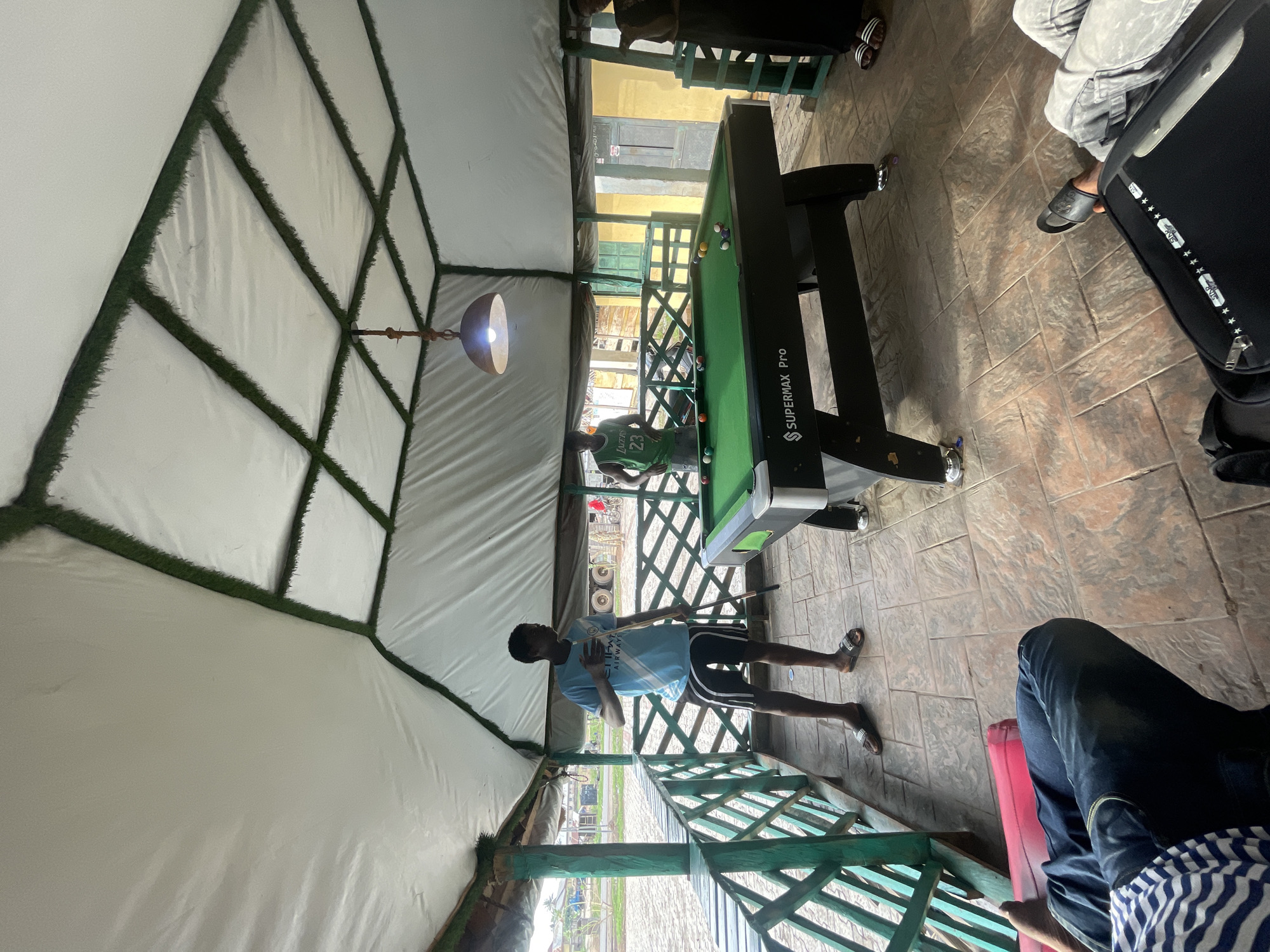 The image depicts an indoor/outdoor recreational space, likely in Lokoja, Nigeria, given the request. The space is characterized by a partially enclosed structure with a white fabric ceiling supported by a geometric frame lined with green artificial turf. The floor is tiled with a textured, stone-like pattern.
A central element is a pool table labeled "SUPERMAX Pro," positioned in the middle of the space. Two young men are present near the table. One is actively playing pool, wearing a Lakers #23 jersey, while the other is standing and holding a pool cue, clad in a light blue Manchester City jersey and black shorts.
Other individuals are partially visible, including a person seated to the side. The foreground shows a seated person wearing jeans and a striped shirt.
A decorative lamp hangs from the ceiling above the pool table. The perimeter of the structure is lined with a green wooden railing. Outside the railing, a glimpse of the Lokoja cityscape is visible.