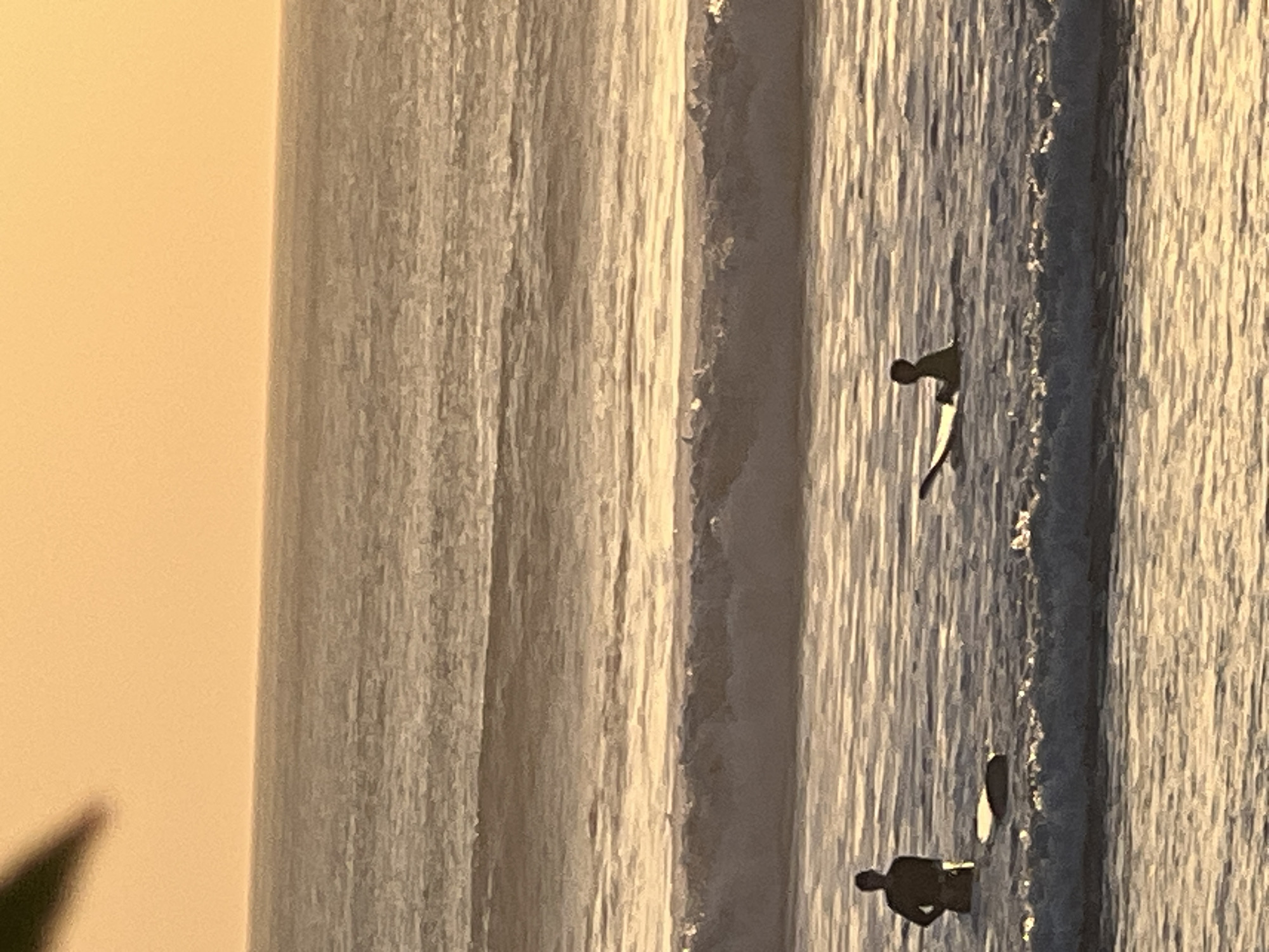 The image depicts a serene ocean scene at sunset in Kuta, Indonesia. Two figures are visible in the water, each with a surfboard. The figure on the left stands partially submerged, facing the viewer. The figure on the right is lying prone on their surfboard, facing away from the viewer. The ocean is calm with gentle waves, and the sky is a warm orange hue, indicating the sun is setting or rising. The overall impression is one of tranquility and outdoor recreation.