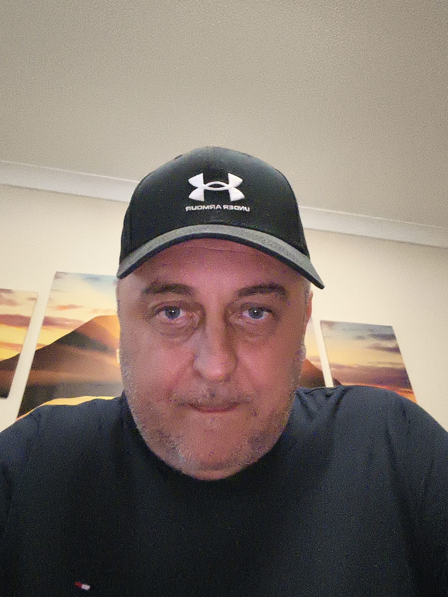 A close-up, eye-level selfie shows a man of middle age, possibly in his 50s, looking directly at the camera. He has a light skin tone, blue eyes, and short, light-colored stubble on his face. His expression is neutral to slightly serious.
He is wearing a black baseball cap with a grey brim edge. The cap features a white Under Armour logo, with the brand name "UNDER ARMOUR" printed below it in white text. The text appears mirrored, reading "RUOMRA REDNU," suggesting the photo was taken with a front-facing camera without image inversion. He is also wearing a dark, likely black or navy, t-shirt. A small, multi-colored emblem, possibly red, white, and blue, is visible on the left chest area of his shirt.
The background consists of a light-colored, textured wall (beige or off-white). On the wall, three panels of artwork are visible. These panels depict a mountain landscape with a vibrant sky in warm hues of orange, red, and purple, suggesting either a sunrise or sunset. The panels are evenly spaced behind the man.
The lighting appears to be artificial, likely overhead, creating some highlights on the man's forehead and nose. The image has a slightly grainy quality. The overall scene suggests an indoor setting within a residence in Huntingdonshire, United Kingdom.