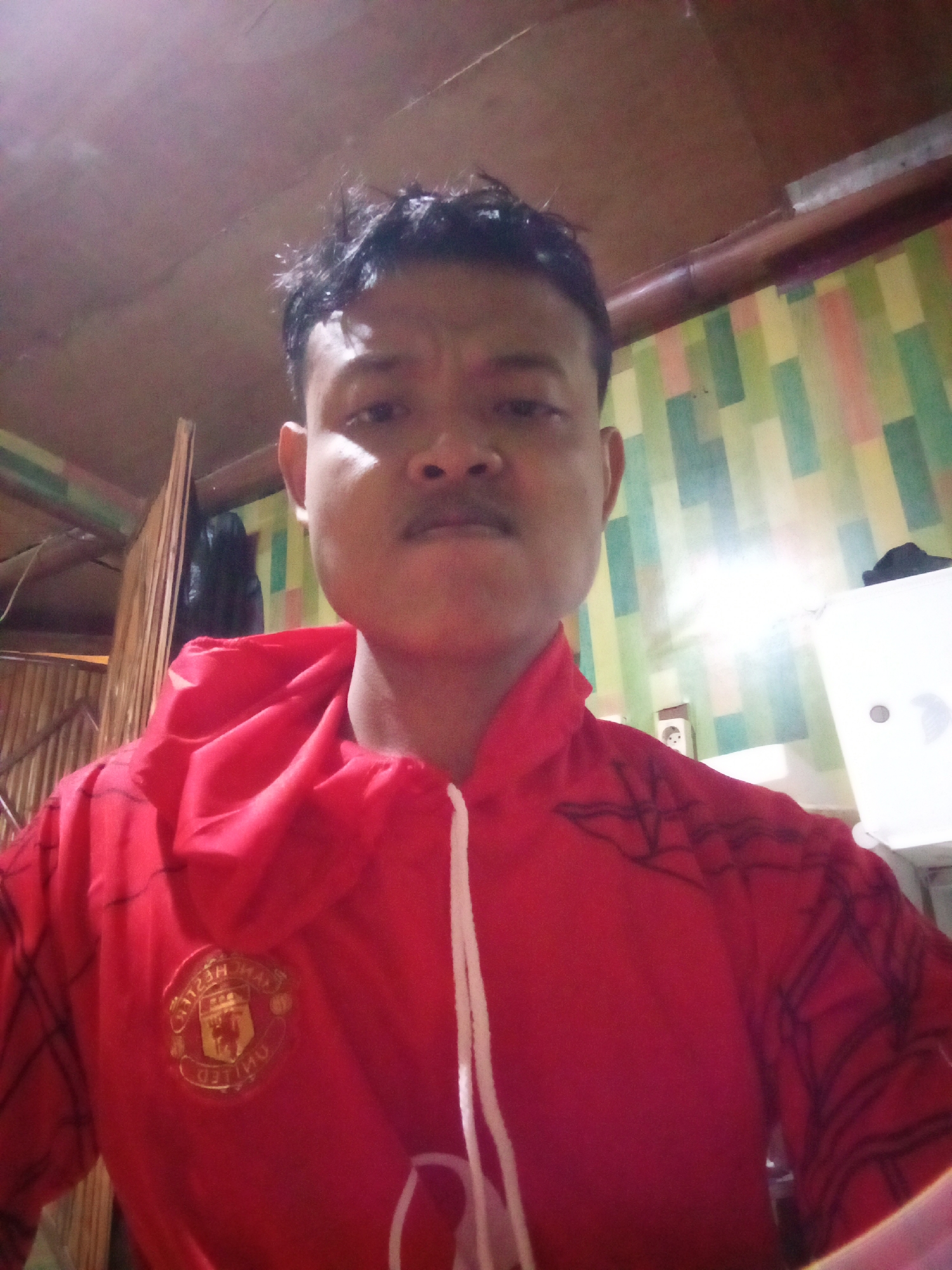 The image shows a young man in a red hooded jacket with a Manchester United logo on the left chest. He is looking directly at the camera with a neutral expression. The background consists of a wall with a geometric pattern of green, yellow, and red rectangles, and a ceiling with wooden beams. A bamboo wall structure is visible on the left side of the image. The lighting is dim, suggesting an indoor setting. The man is wearing the jacket zipped up, with white drawstrings visible at the neckline. The scene appears to be a selfie taken in a domestic environment. While the image does not explicitly depict Candi Wates, Indonesia, the user has specified this location.