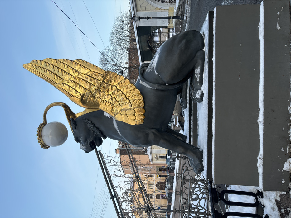 The image displays one of the iconic winged griffin statues from the Bank Bridge (Bankovsky Bridge) in Saint Petersburg, Russia. The griffin, cast in a dark, possibly bronze material, is positioned prominently on a dark gray rectangular pedestal lightly dusted with snow. Its large, feathered wings are gilded in a striking gold and extend upwards. A spherical white lamp, topped with a golden crown-like ornament, protrudes from the griffin's back via a curved golden arm. A light layer of snow is visible on the griffin's back, legs, and the top surfaces of the pedestal.
To the immediate left, an ornate dark metal railing, part of the bridge, is partially visible, also bearing traces of snow. In the background, across a visible waterway or canal, classical-style multi-story buildings with facades in light yellow and orange hues line a street where several cars are parked. Overhead, a network of wires supports numerous small decorative lights, possibly spanning the bridge. To the right of the griffin, a dark street extends into the distance, bordered by a traditional black lamppost and several bare deciduous trees. The scene is illuminated by a clear, light blue winter sky. No people are present in the image.