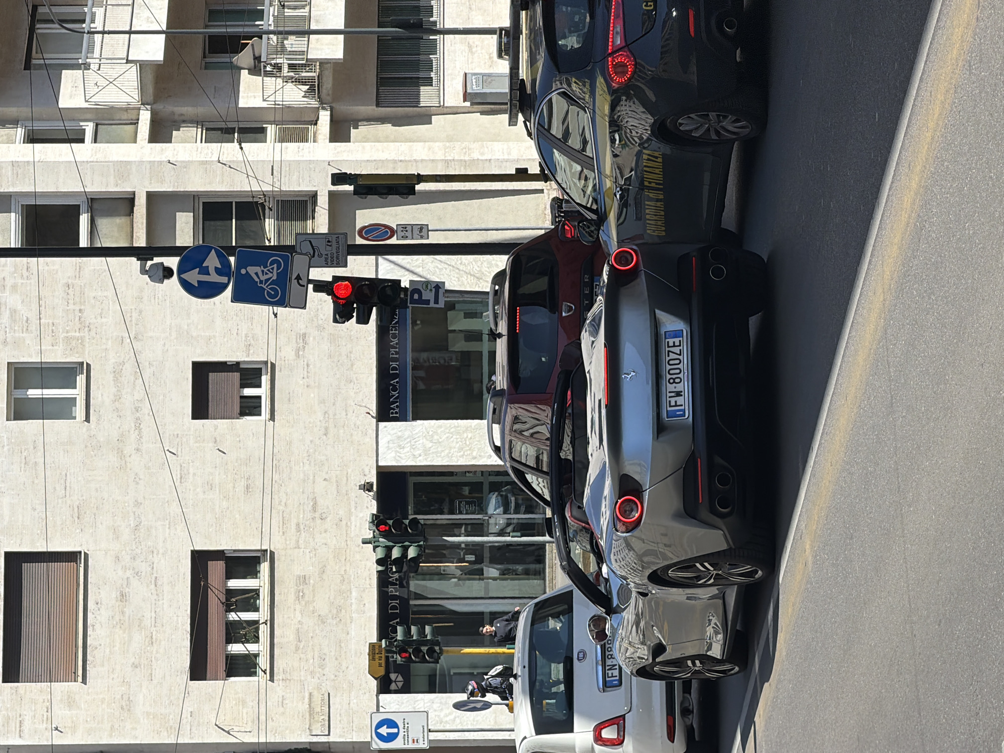The image shows a street scene in Milan, Italy. The location is identifiable due to the presence of the Banca di Piacenza building and a sign indicating "Porta Vittoria." The street is congested with parked cars. Among them is a grey sports car with license plate "FW-800ZE" and a car with "GUARDIA DI FINANZA" markings, indicating it is part of the Italian Financial Police. Various traffic signs are visible, including a directional sign for cyclists and a surveillance sign. A man is standing by one of the cars near the bank. The traffic lights are red. The buildings are typical of Italian urban architecture.
