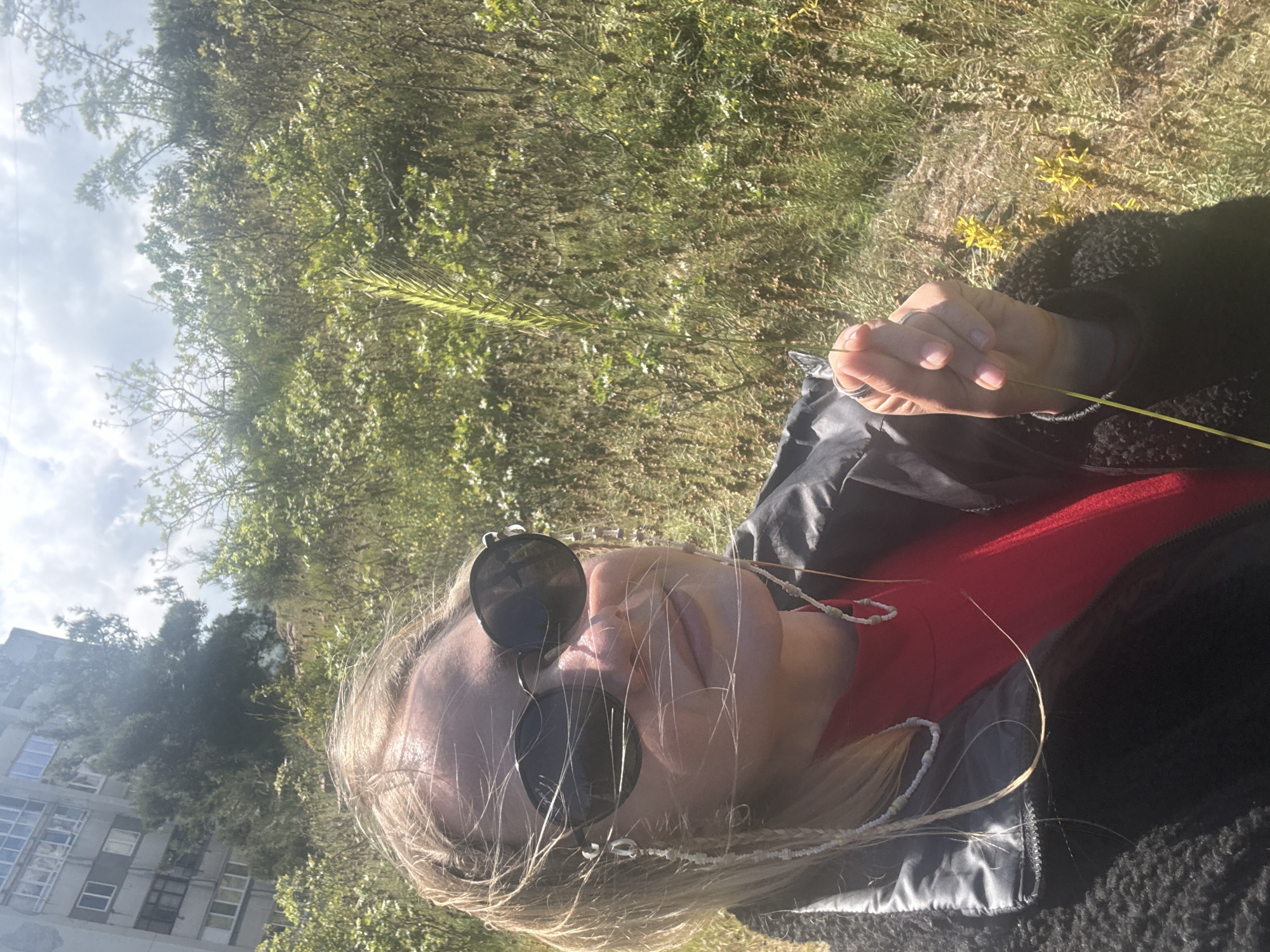 A low-angle, medium shot captures a person wearing sunglasses and a red shirt, with a black jacket, holding a blade of grass. The background features green foliage, suggesting a natural setting. In the lower left, a building is partially visible. The location appears to be outdoors, with a mix of natural and urban elements.