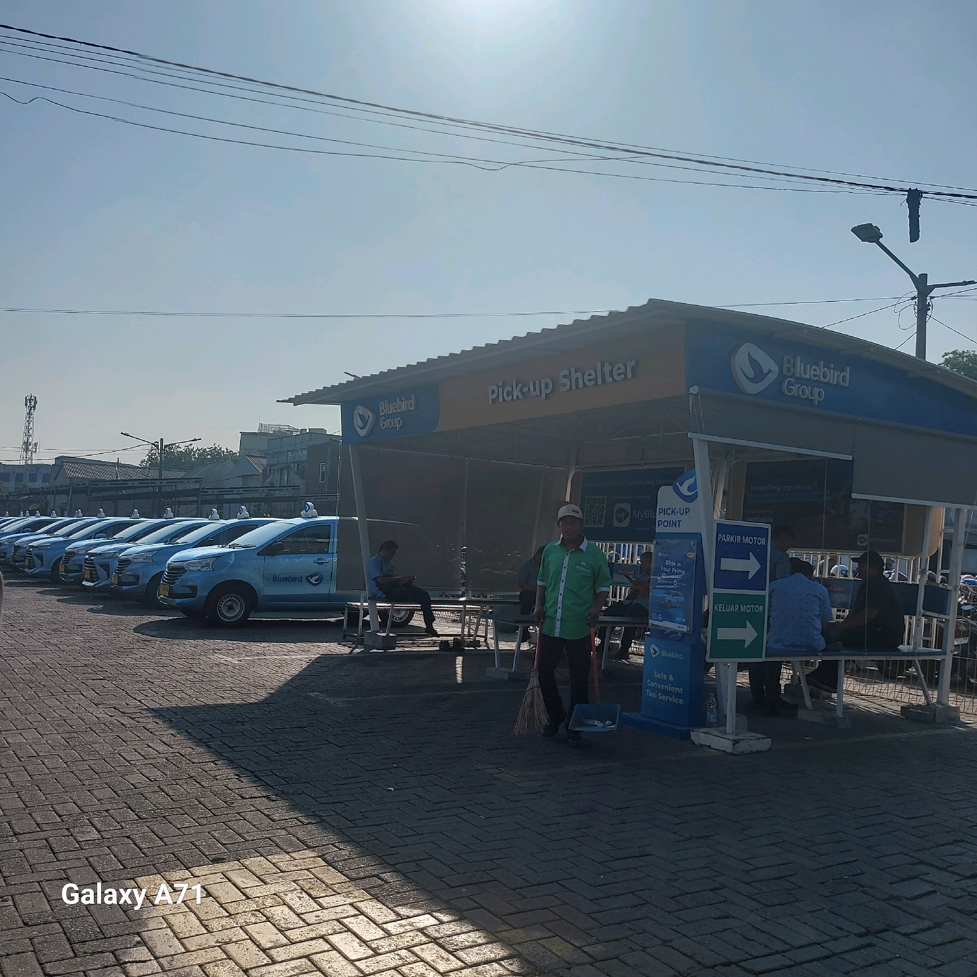 In Surabaya, Indonesia, a Bluebird Group "Pick-up Shelter" is prominently featured, situated adjacent to a row of parked Bluebird taxis. The shelter, adorned with the Bluebird logo, also displays signage including "Pick-up Shelter," "MyBluebird PICK-UP POINT," and directions for "PARKIR MOTOR" (Motorcycle Parking) and "KELUAR MOTOR" (Motorcycle Exit).
A man in a green shirt and dark pants stands near the shelter, holding a broom and dustpan, suggesting he is performing cleaning duties. Several other individuals are visible in and around the shelter, some seated on benches and others standing. The taxis are light blue with the "Bluebird" logo. The ground is paved with dark grey interlocking bricks, casting shadows from the shelter and a nearby light pole. In the background, buildings and a cell tower are visible under a bright, hazy sky. The scene suggests an active taxi service hub operating during daylight hours.