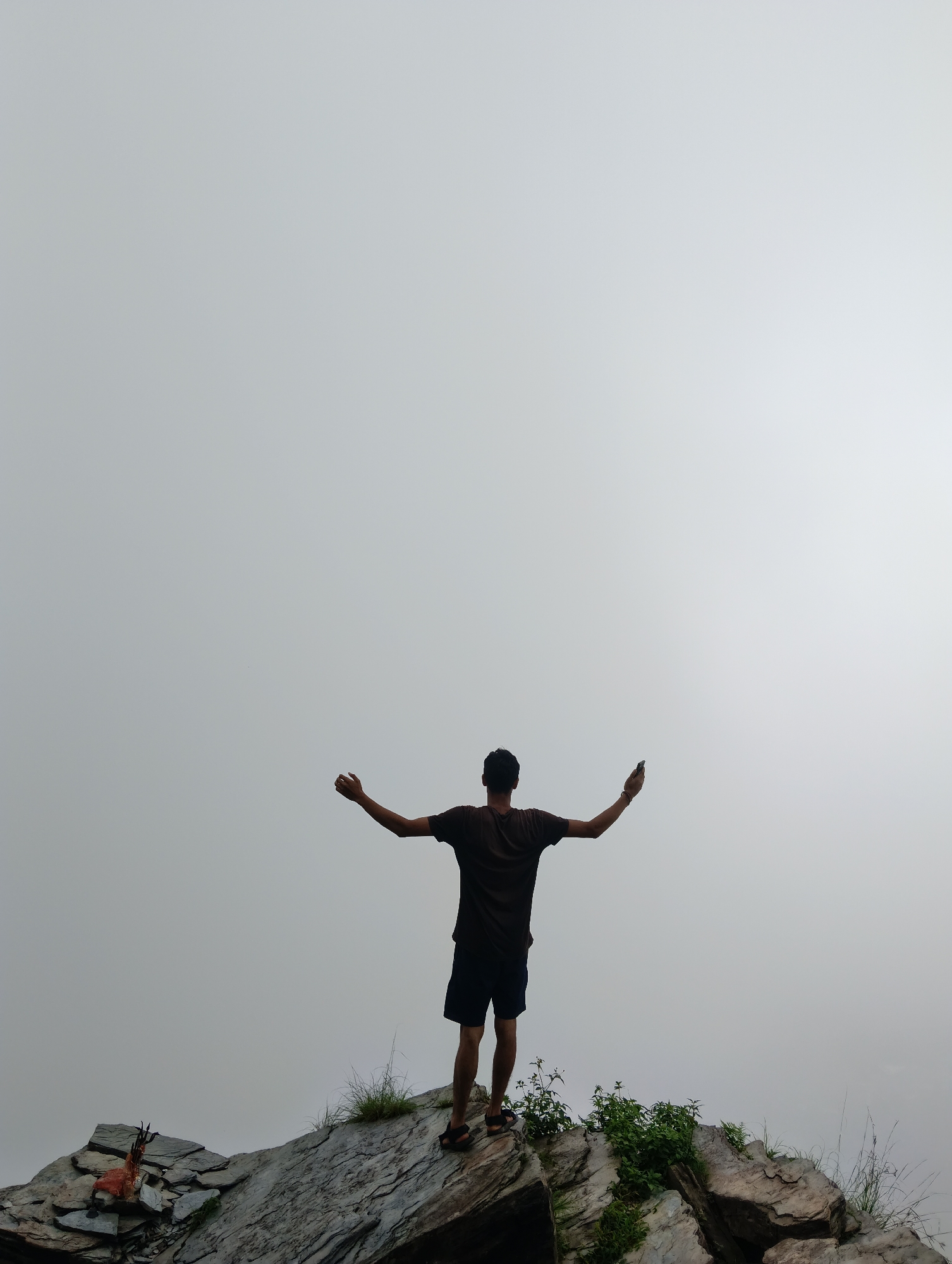 In Bhagsu, India, a person stands on a rocky outcrop with their arms outstretched, silhouetted against a light gray sky, possibly indicating overcast conditions or fog. The individual, wearing a dark t-shirt and shorts, is facing away from the camera and holding a mobile phone in their right hand. In the lower left of the frame, a small statue or effigy, possibly of a goat, rests on a pile of flat stones. The rocky terrain is covered with sparse green vegetation.
